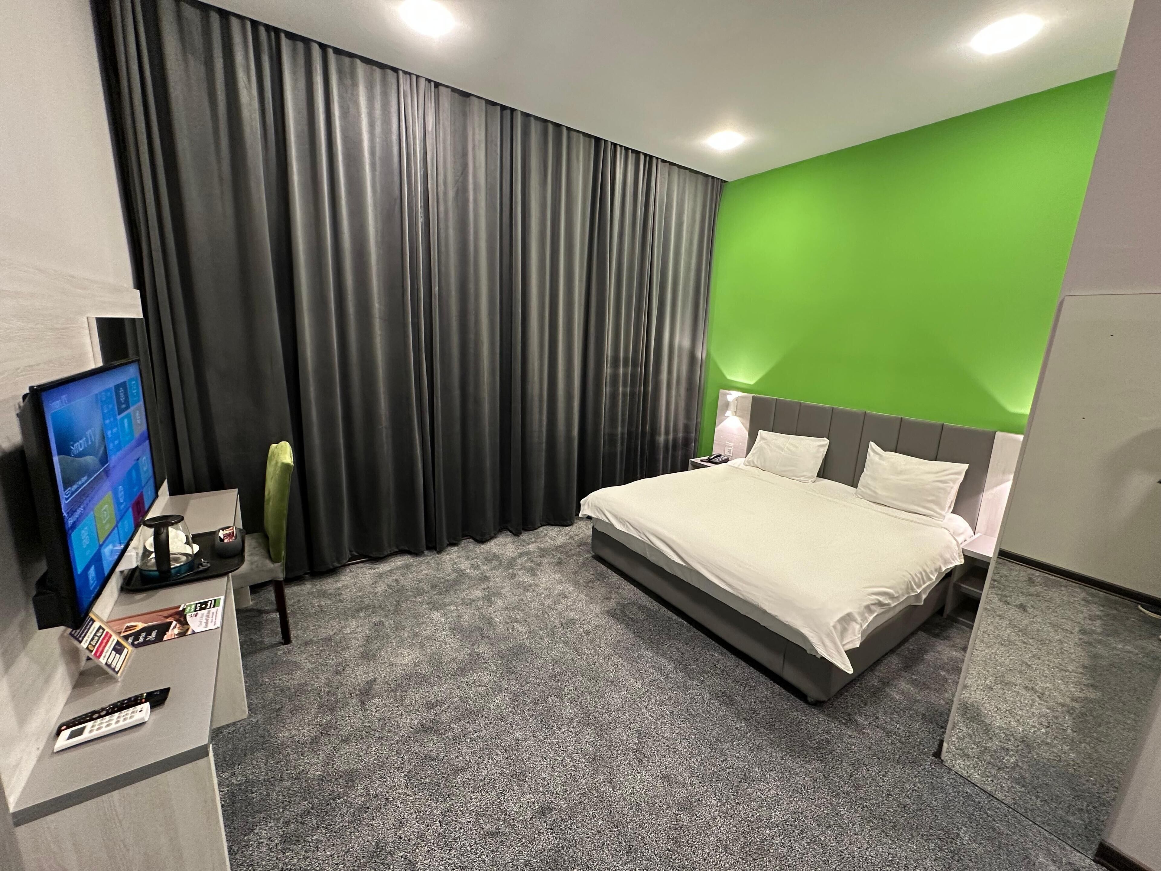 A modern hotel room with a large bed, a flat-screen TV, a desk with a lamp, and a green accent wall.