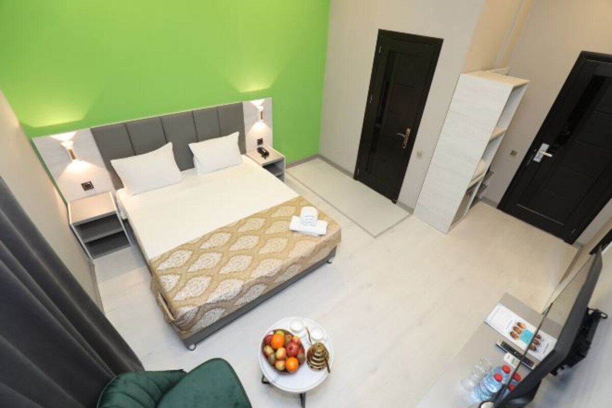 A hotel room with a bed, bedside tables, a green wall, and a small table with fruit and a bottle.