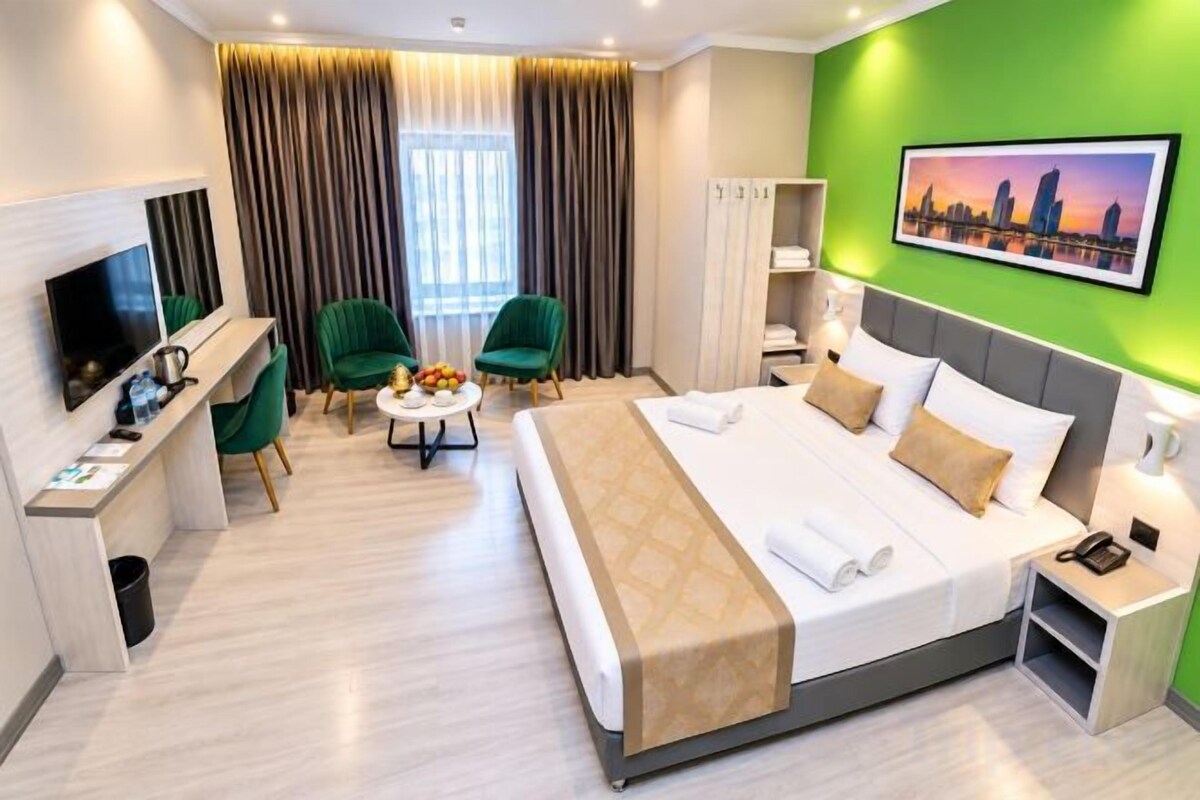 A hotel room with a bed, a TV, a desk, a chair, a small table, and a painting on the wall.