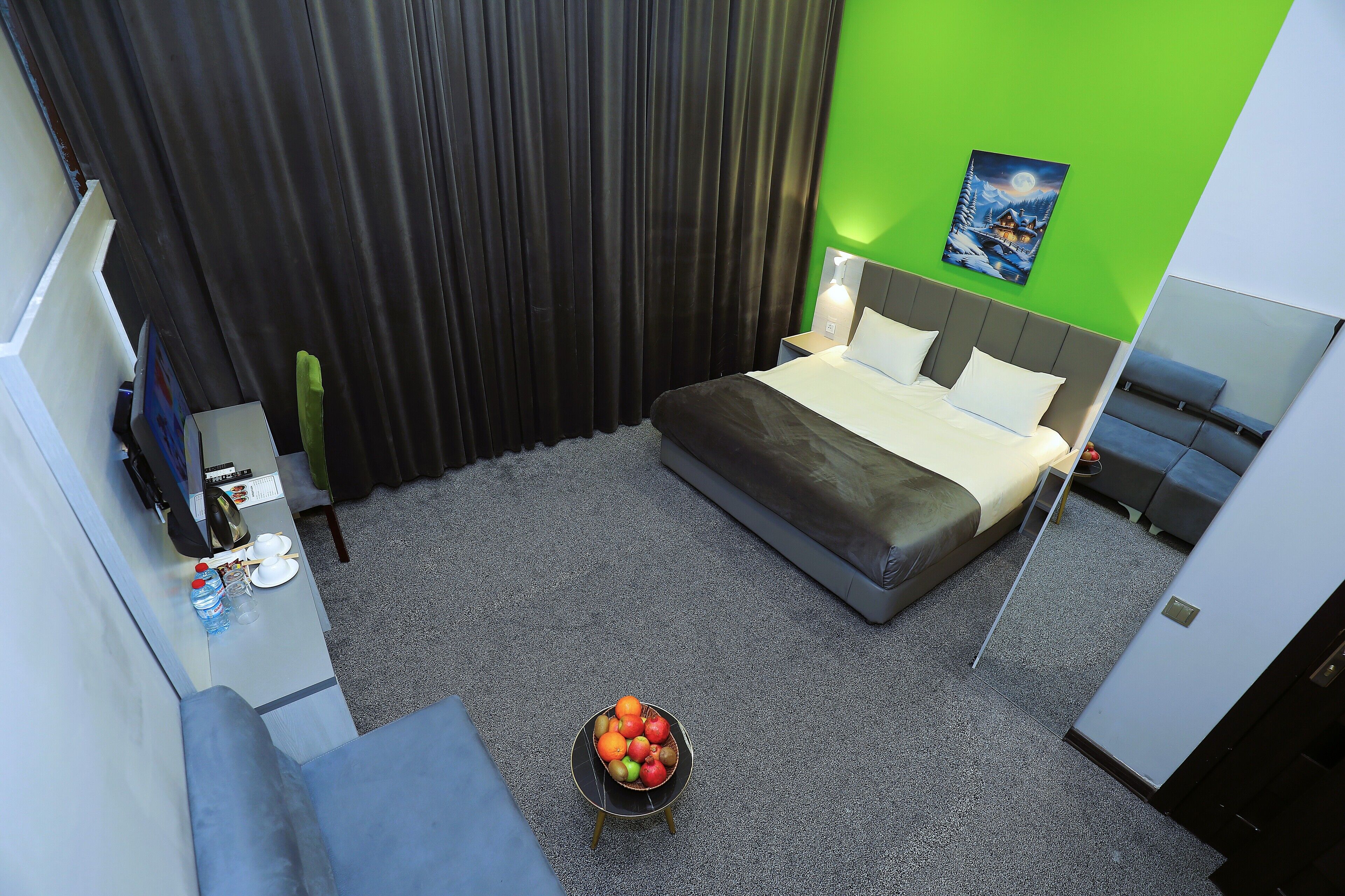 A hotel room with a bed, a small table with a fruit bowl, a nightstand, a TV, and a green wall.