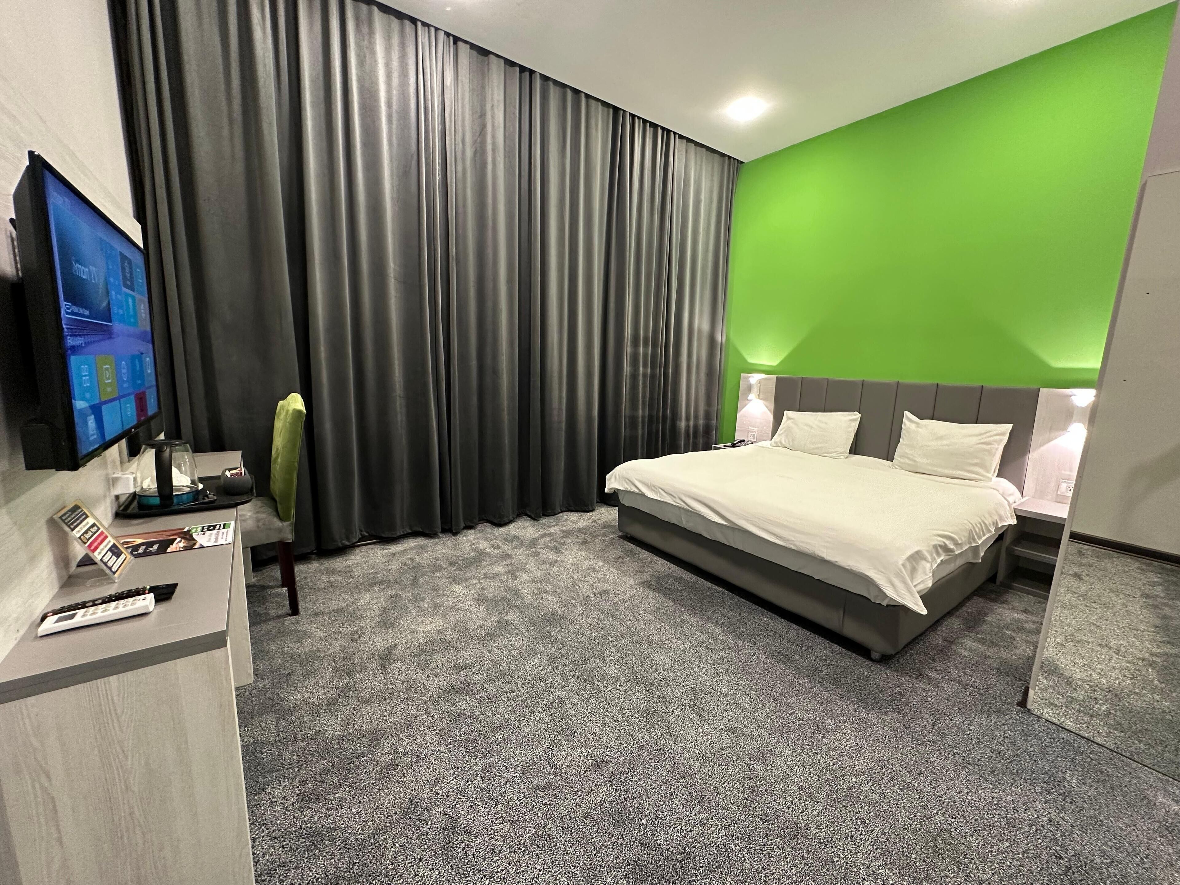 A modern hotel room with a large bed, a desk with a computer and remote controls, a television, and a green accent wall.