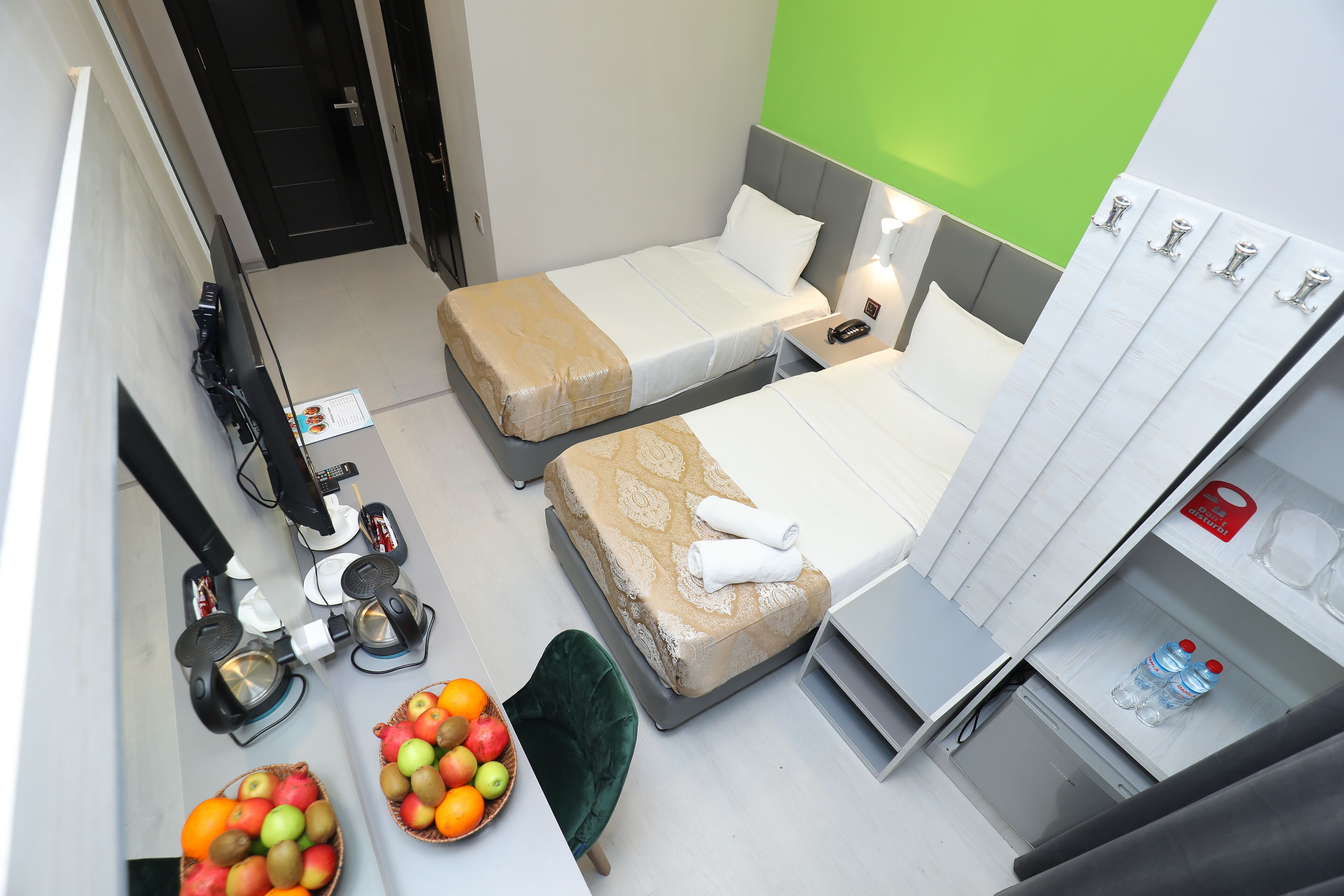 Ein Hotelzimmer mit zwei Betten, einem Schreibtisch mit Obstschale, einem grünen Sessel, einer Minibar und einer grünen Wand.