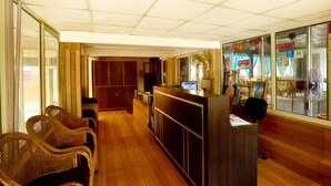 A reception area with a wooden reception desk, a computer, and a glass display case with various items. There are wicker chairs and a wooden cabinet in the background. - Kunwar Residency (Narendranagar)