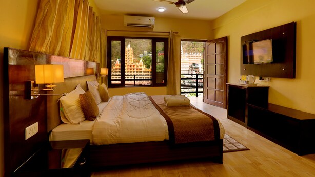 A hotel room with a bed, a TV, an air conditioning unit, and a view of a temple through the window. - Kunwar Residency (Narendranagar)