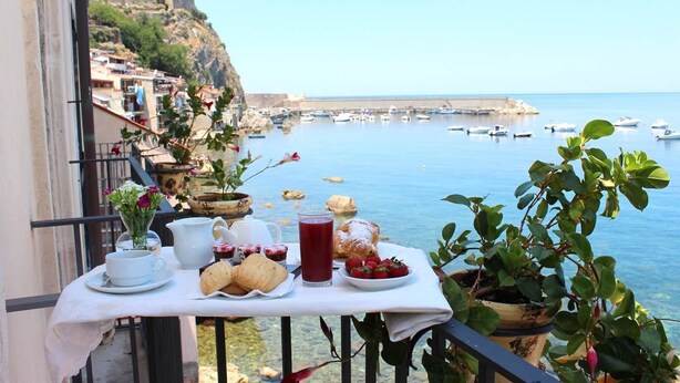 Un balcon avec une table préparée pour le petit-déjeuner, donnant sur une ville côtière avec des bateaux et un fort historique.