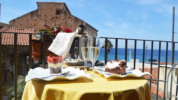 A table set with a dessert, two glasses of white wine, and a glass of water on a balcony with a view of the sea and a historic building. - Hotel Palazzo Krataiis (Scilla)