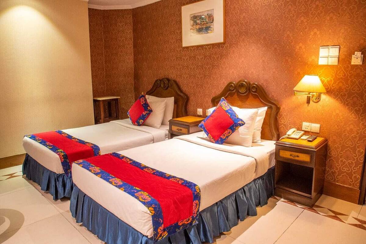 Habitación de hotel con dos camas, cada una con una colcha estampada de flores rojas y azules, cabeceros de madera y una mesita de noche con lámpara y teléfono.