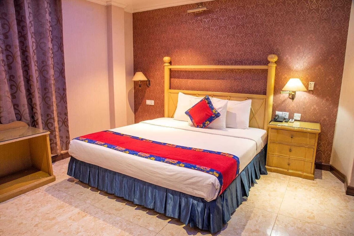 Una habitación de hotel con una cama grande, una mesita de noche con lámpara, un aparador de madera y una cortina.