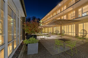 An outdoor patio area with tables and chairs, a large umbrella, and a planter with plants. - Tagungshaus Martin-Niemöller (Schmitten)