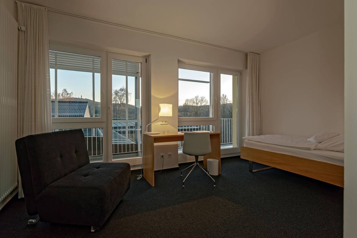 A hotel room with a bed, a desk, a chair, and large windows.