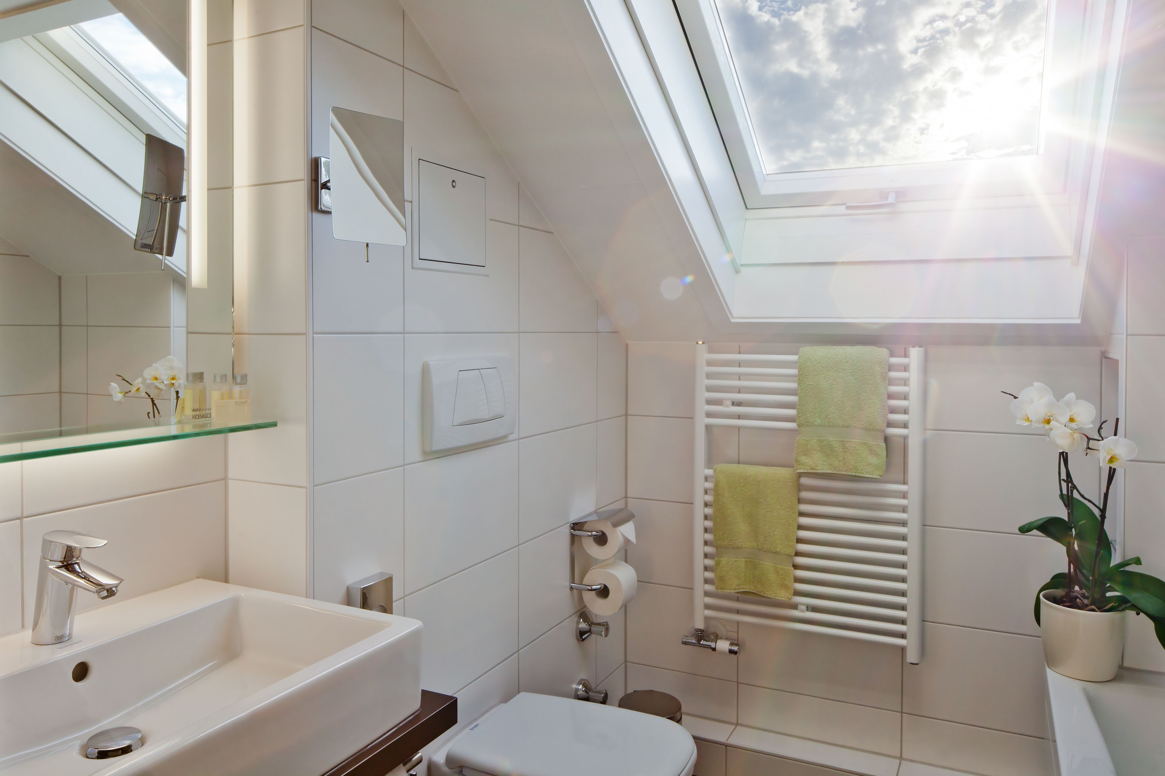 A modern bathroom with a skylight, a white sink, a toilet, a towel rack, and a potted orchid.
