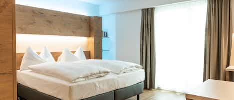 Una habitación de hotel moderna con una cama grande, cabecera de madera, un escritorio y un ventanal con cortinas.