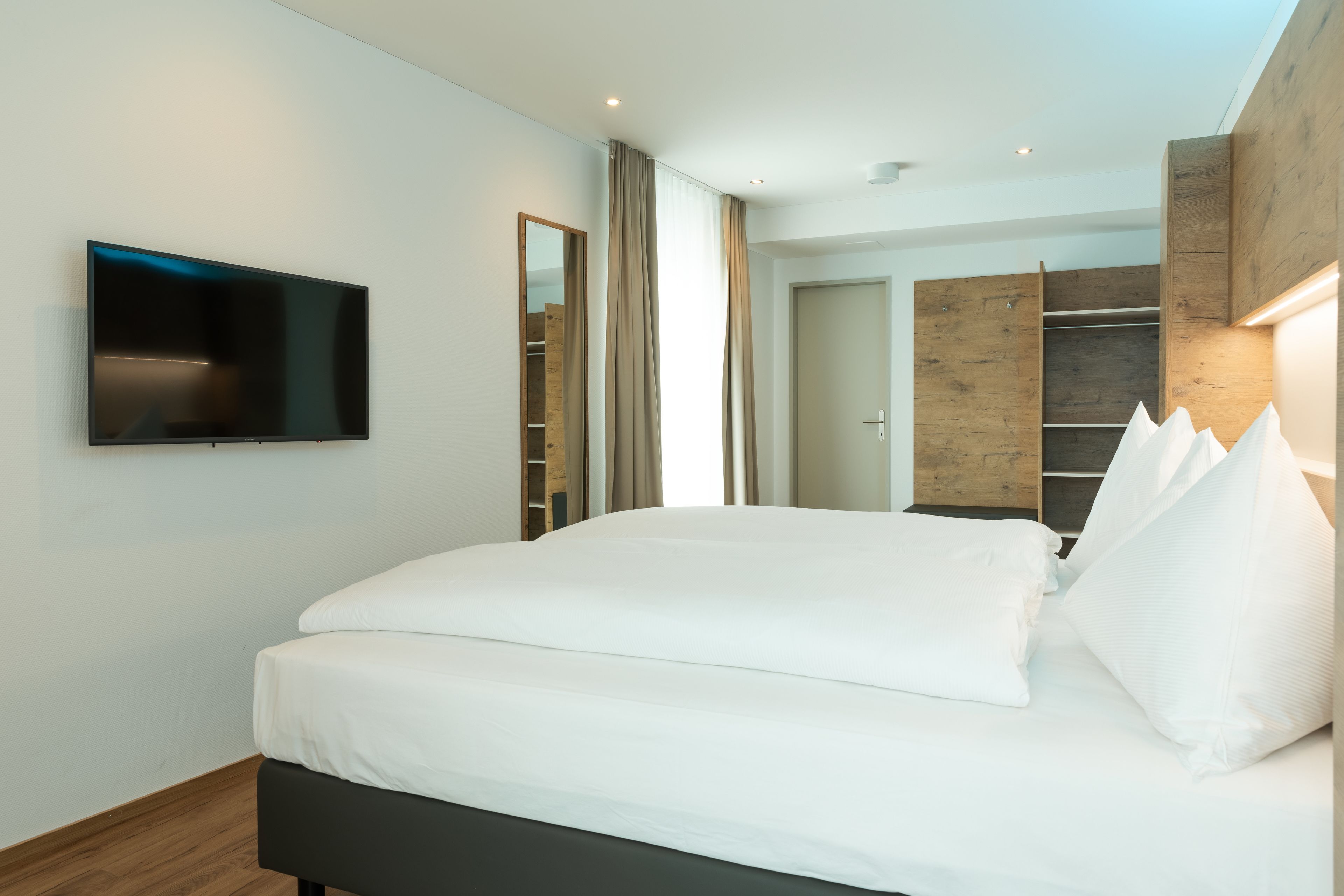 A modern hotel room with a large bed, a flat-screen TV, a wooden wardrobe, and a built-in closet.