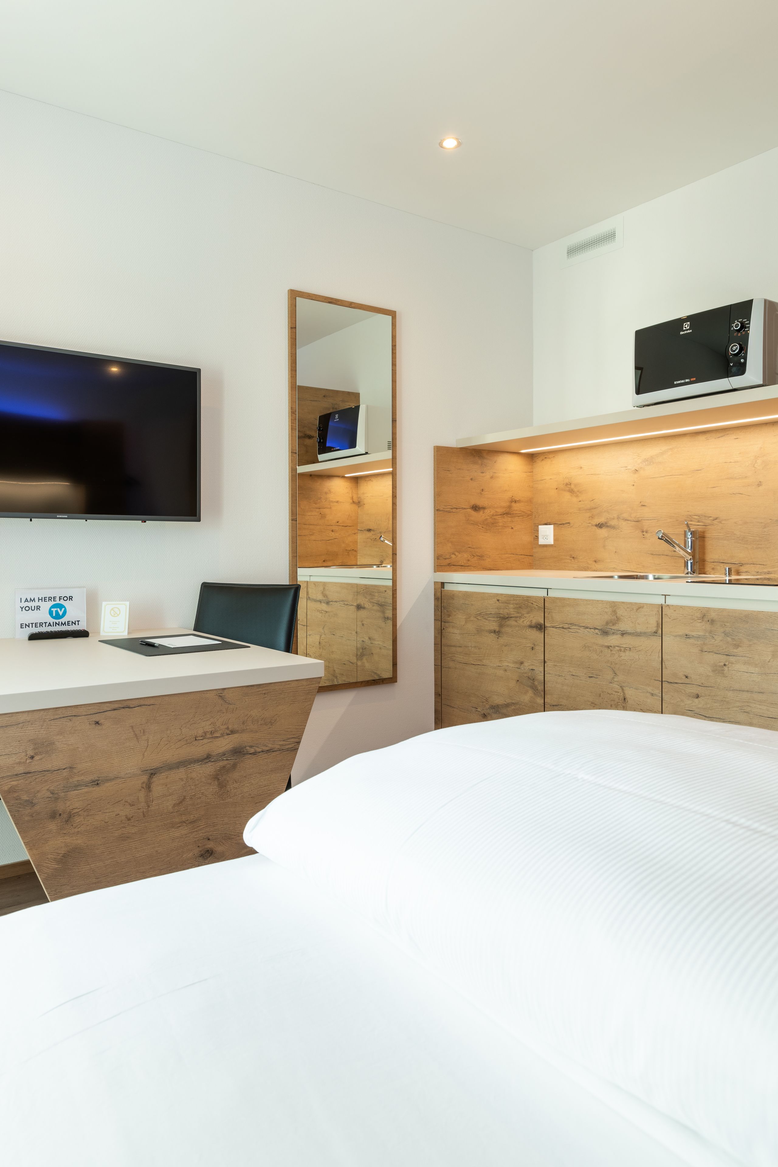 Una camera d'albergo con un letto, una scrivania con un cartello, una TV, uno specchio, un armadietto in legno e un forno a microonde.