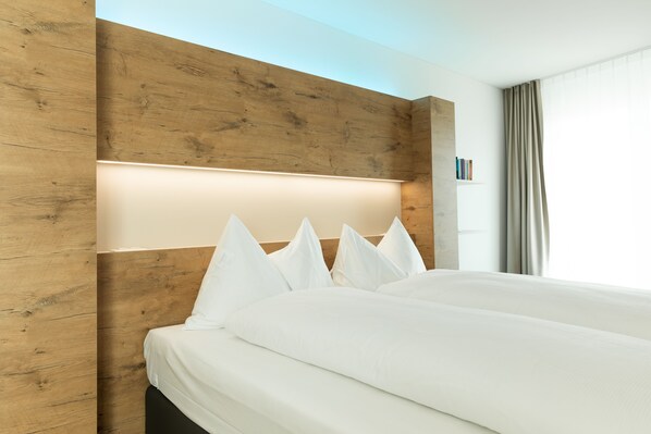 A modern hotel room with a wooden headboard, a neatly made bed with white linens, and a window with curtains.