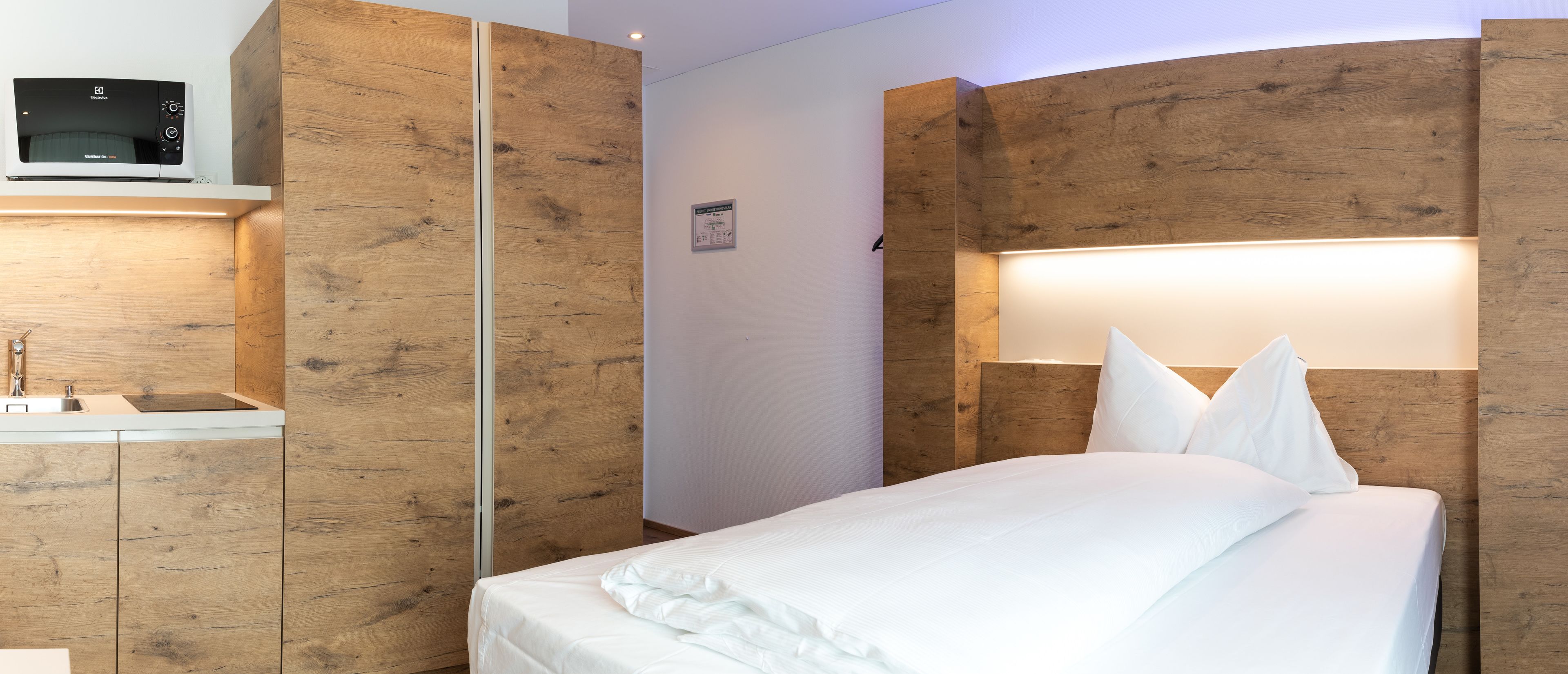 Una habitación de hotel moderna con paneles de madera, una cama con ropa de cama blanca, un microondas integrado y una mesita de noche con lavamanos.