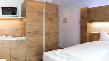 Una habitación de hotel moderna con paneles de madera, una cama con ropa de cama blanca, un microondas integrado y una mesita de noche con lavamanos.