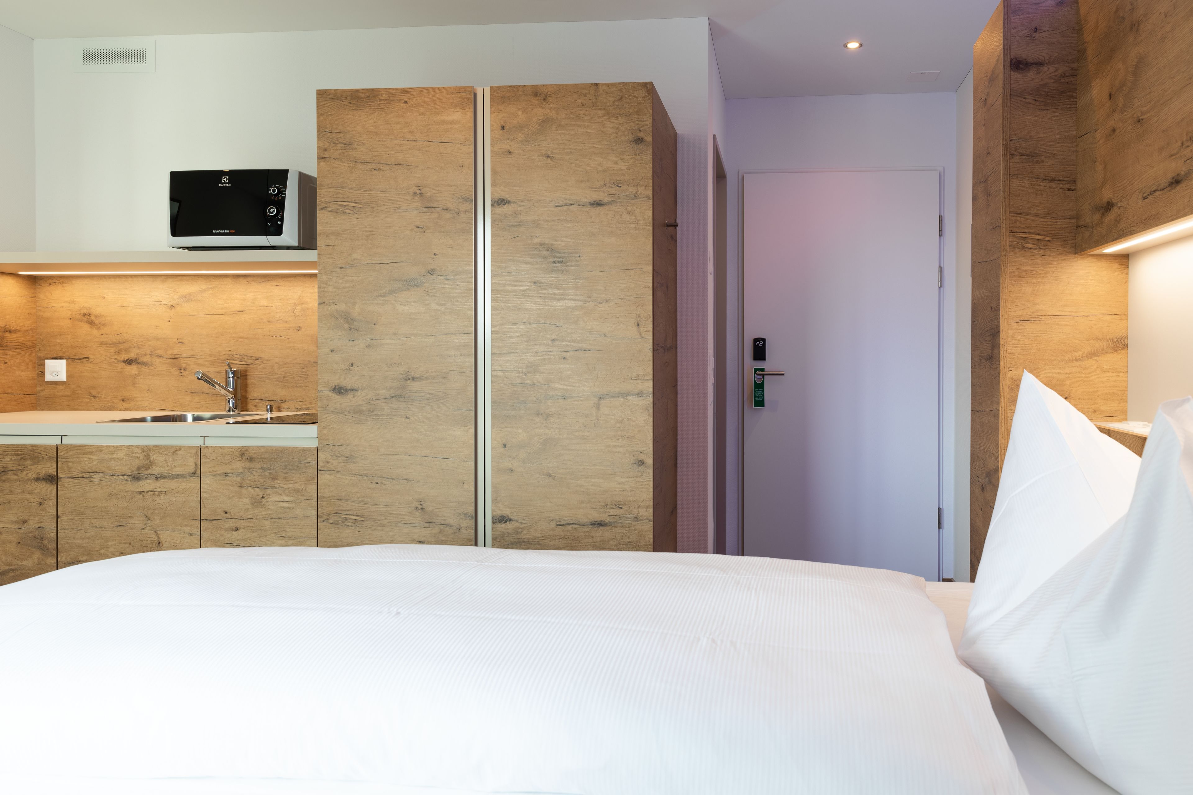 A modern hotel room with a wooden wardrobe, a built-in kitchen with a sink, and a separate door leading to the bathroom.