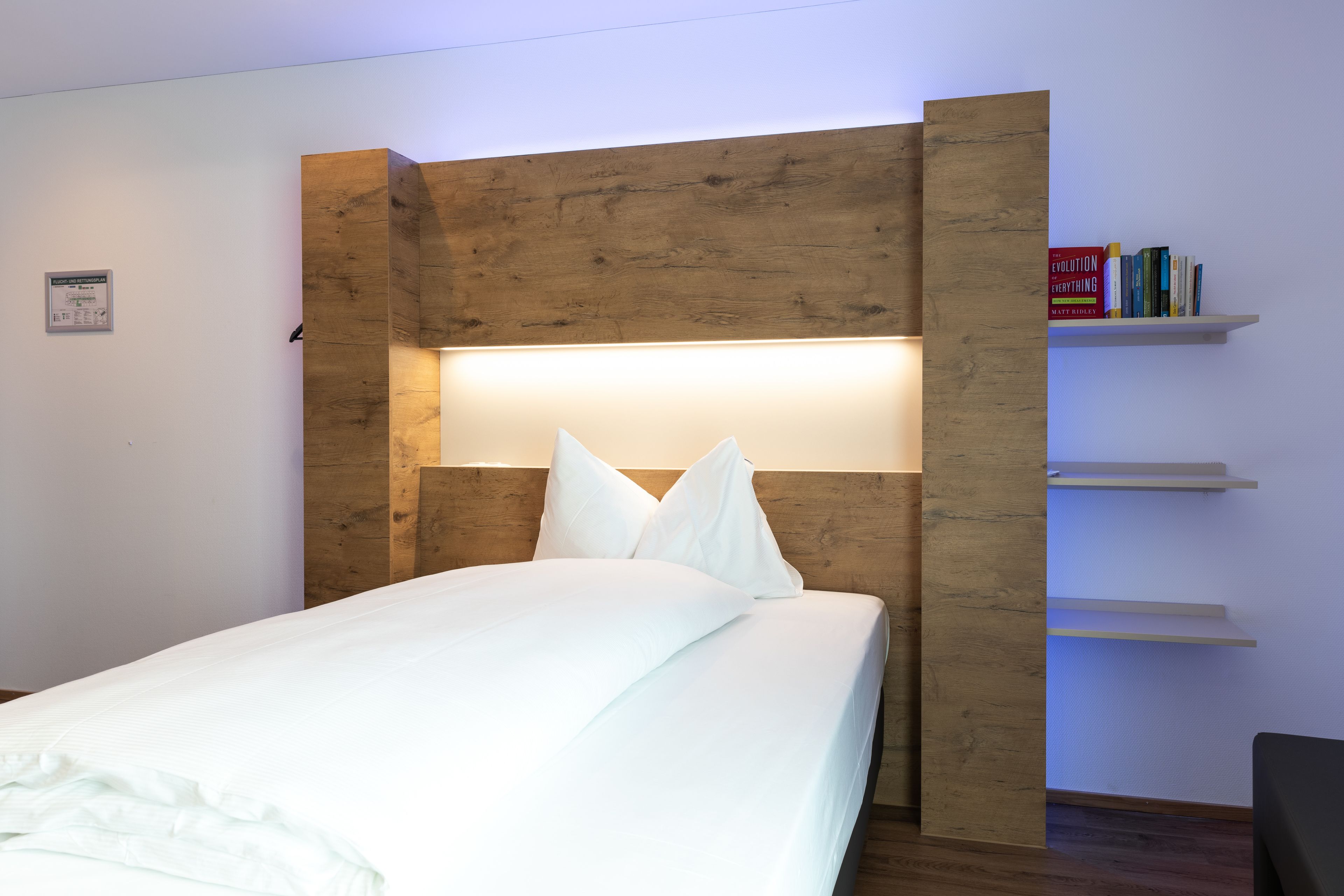 Una cama bien tendida con sábanas y almohadas blancas, un cabecero de madera con iluminación integrada y una estantería con libros a la derecha.