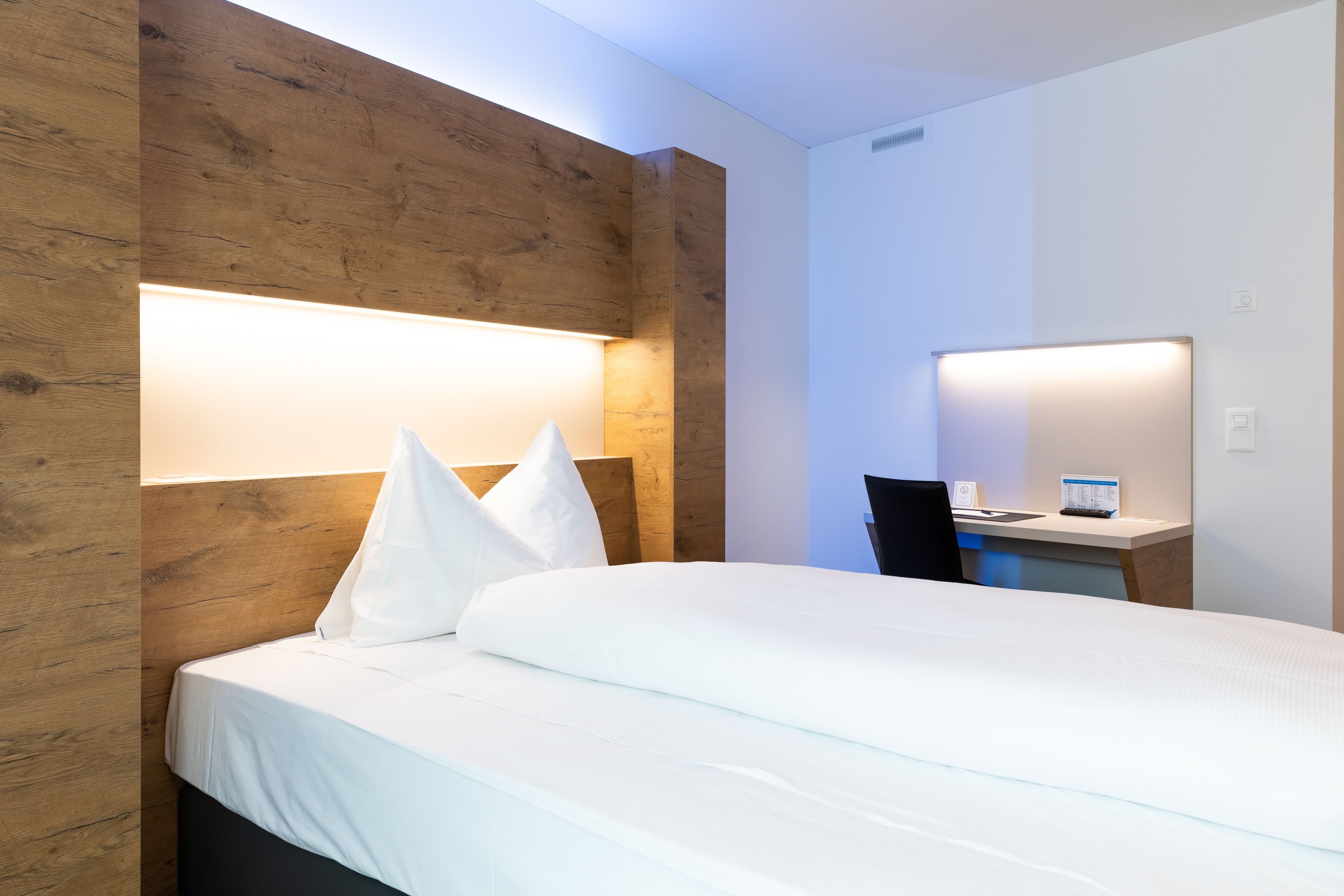 A hotel room with a wooden headboard, a bed with white linens, a desk with a chair, and a wall-mounted lamp.