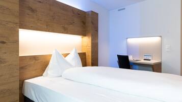 A hotel room with a wooden headboard, a bed with white linens, a desk with a chair, and a wall-mounted lamp.