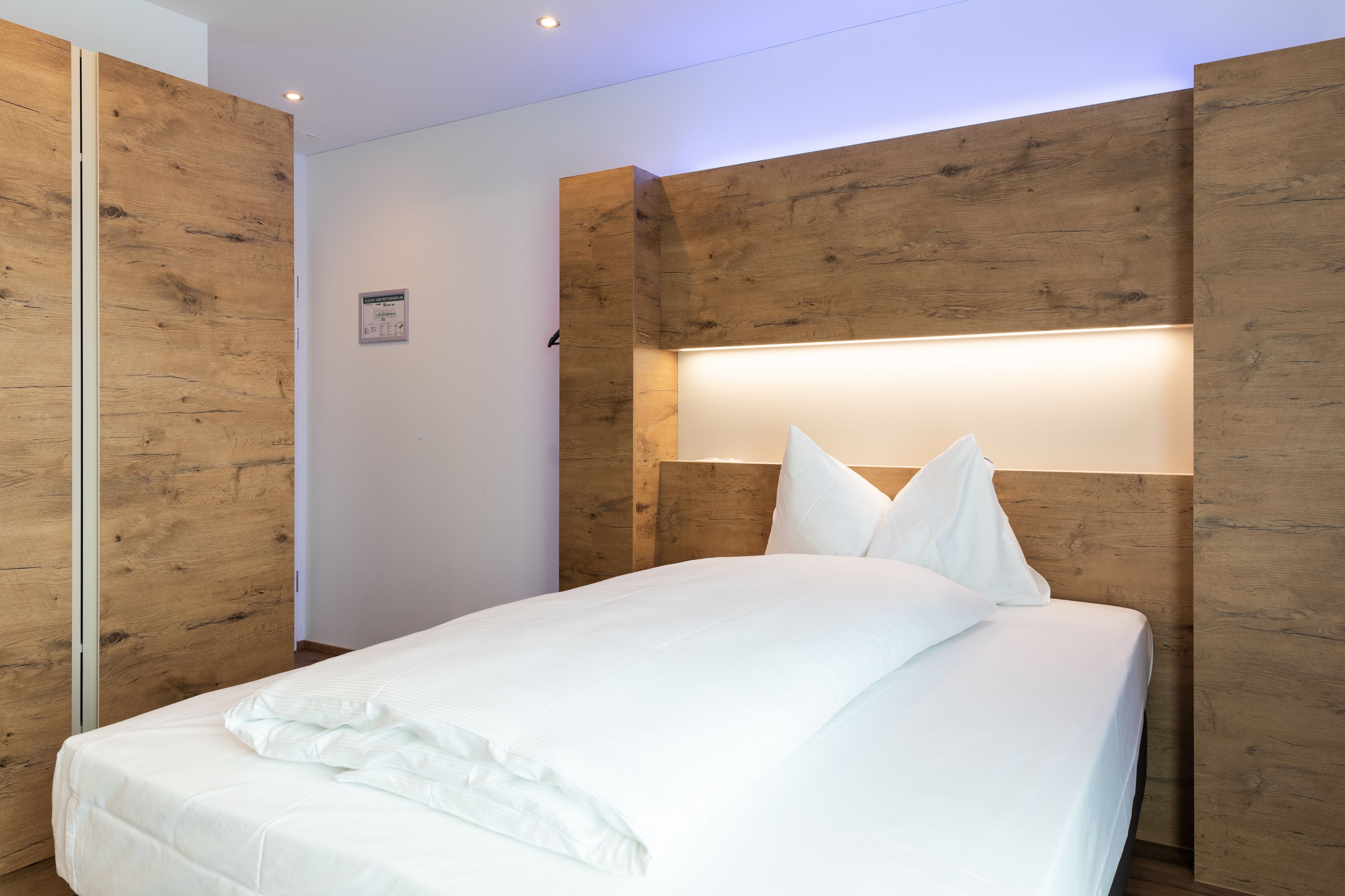 A modern bedroom with a wooden headboard, a neatly made bed, and a wall-mounted light.