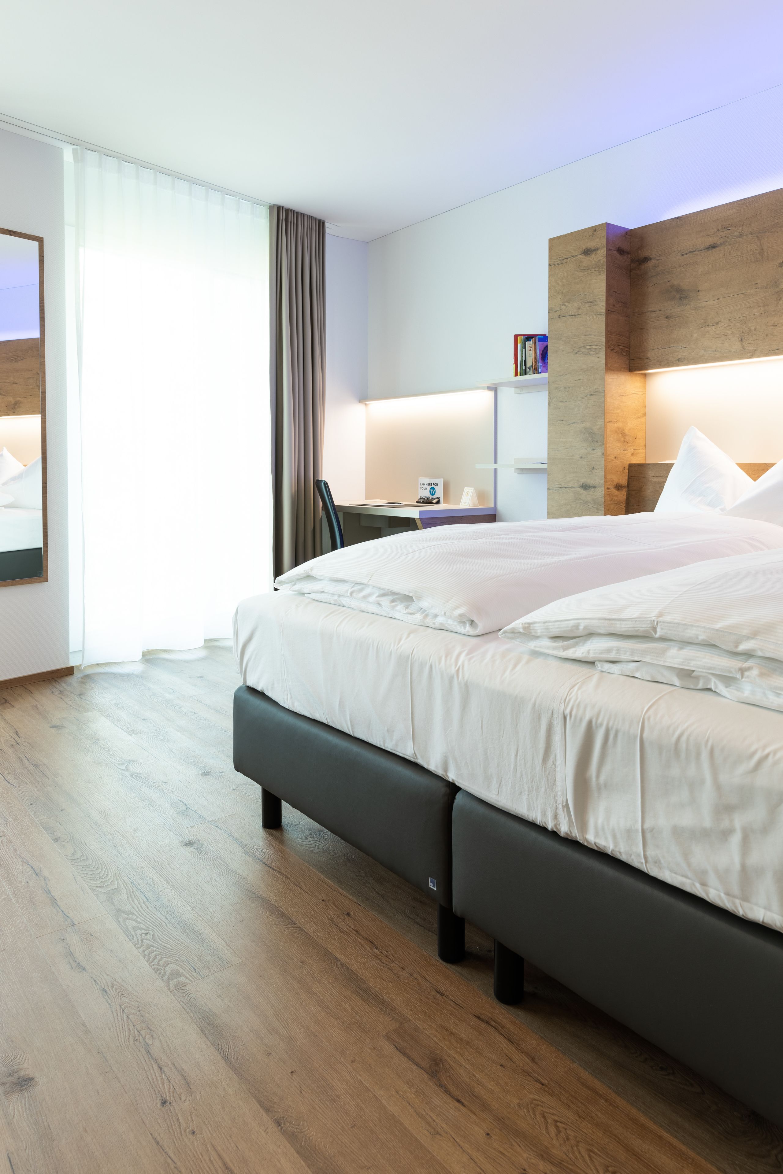 A modern hotel room with a large bed, wooden flooring, a desk with a chair, and a bookshelf.