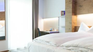 A modern hotel room with a large bed, wooden flooring, a desk with a chair, and a bookshelf.