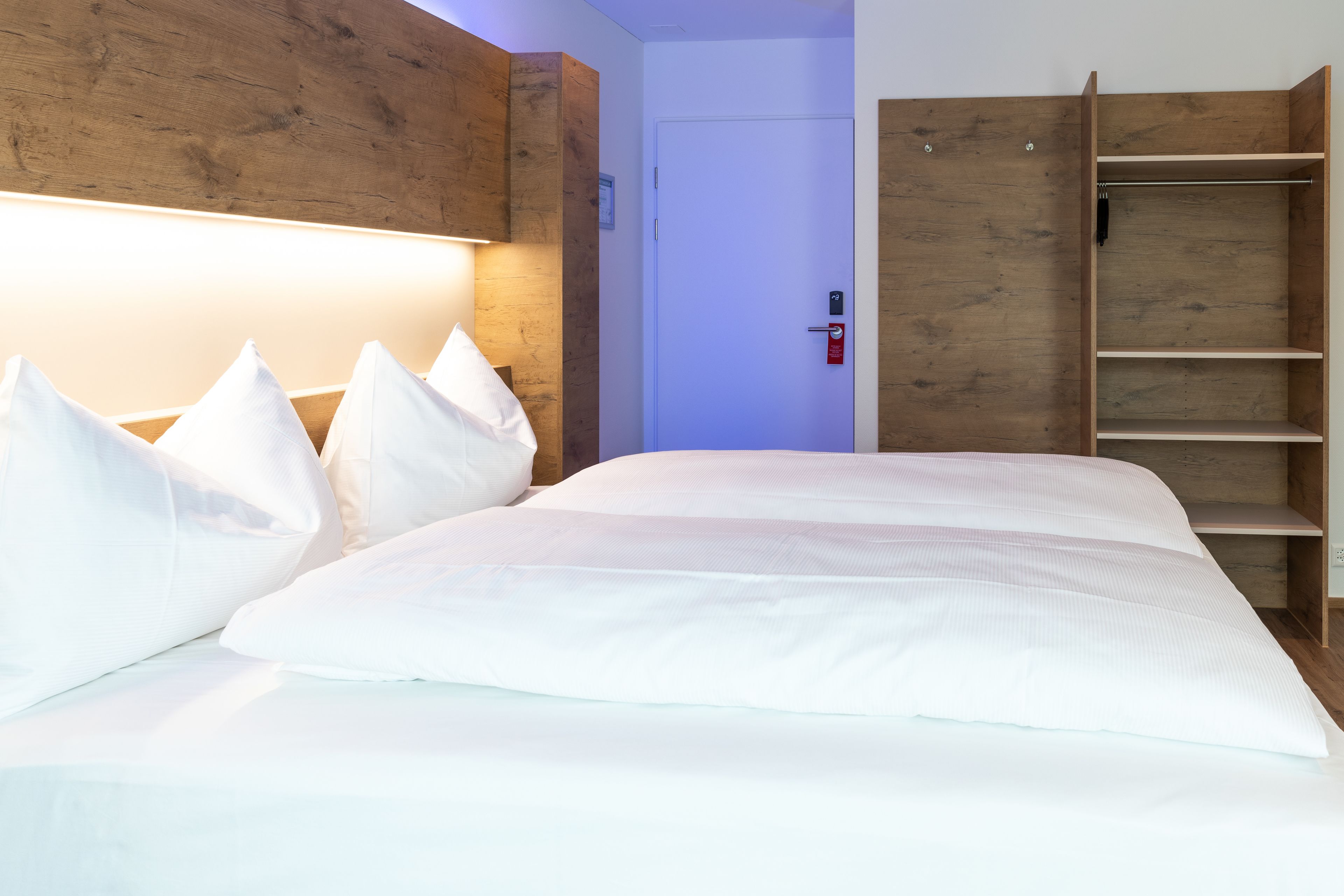 A neatly made bed with white linens and pillows, a wooden headboard, and a wooden wardrobe with open doors.