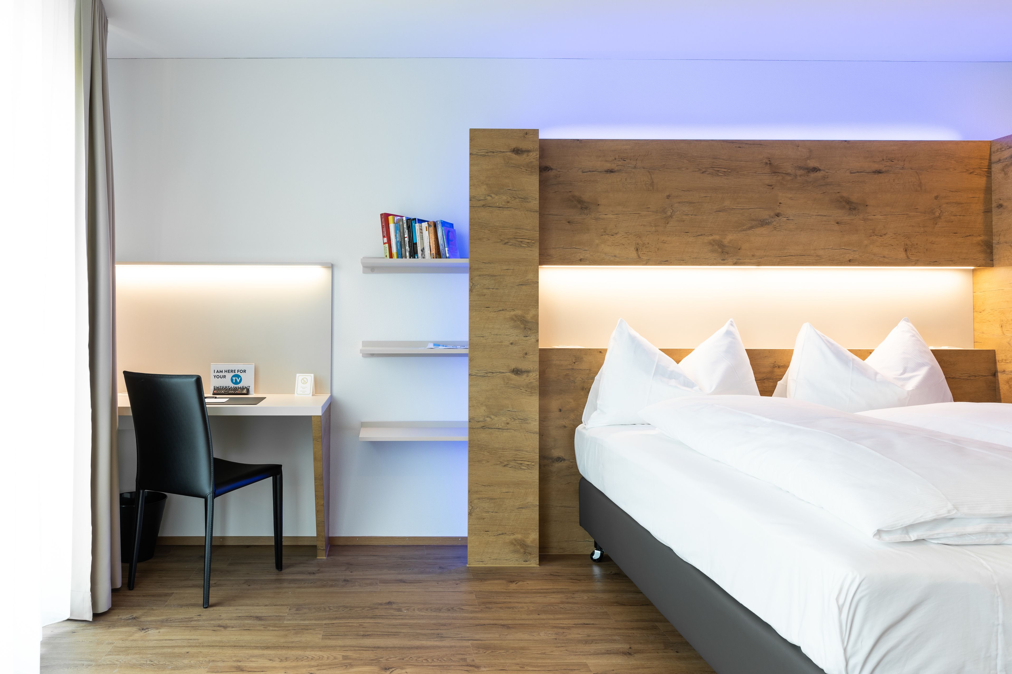 Una habitación de hotel moderna con un cabecero de madera, una cama con ropa blanca, un escritorio con una silla y una estantería con libros.