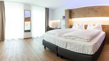 A modern hotel room with a large bed, wooden flooring, a desk with a chair, and a large mirror.