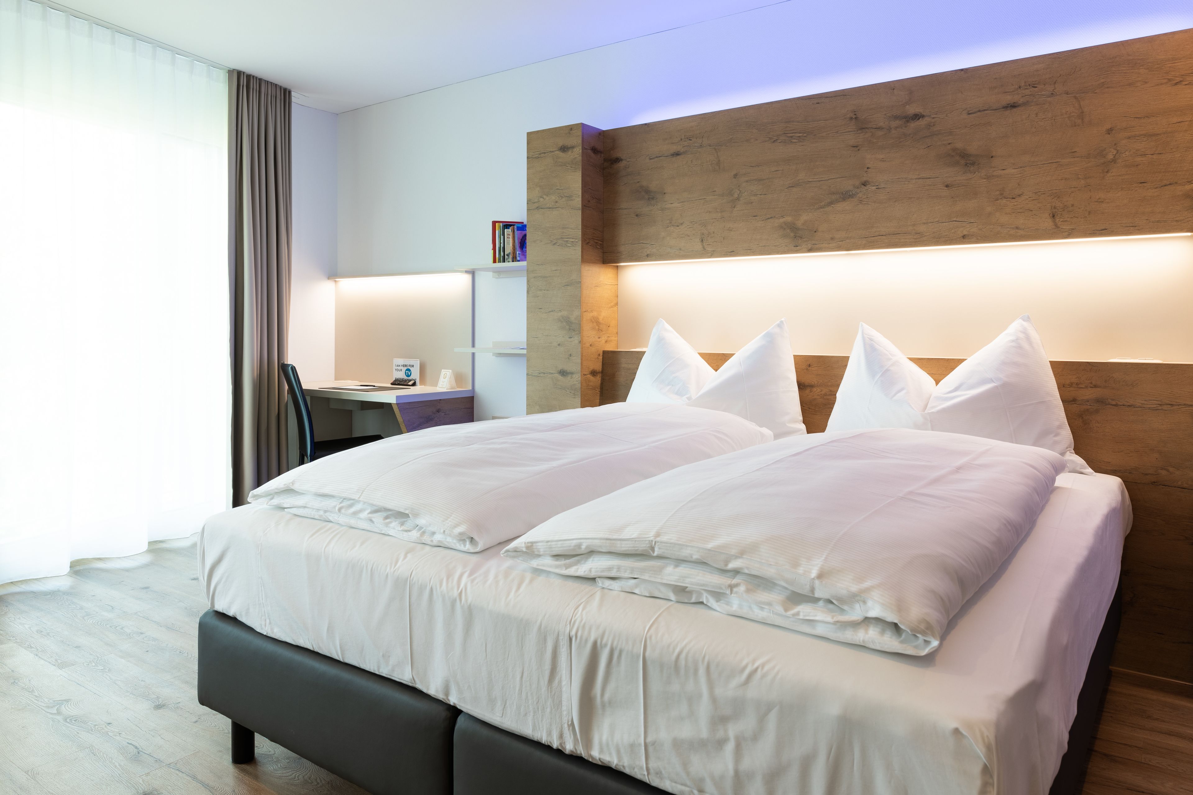 Una habitación de hotel moderna con una cama grande, cabecera de madera, un escritorio con silla y una estantería.