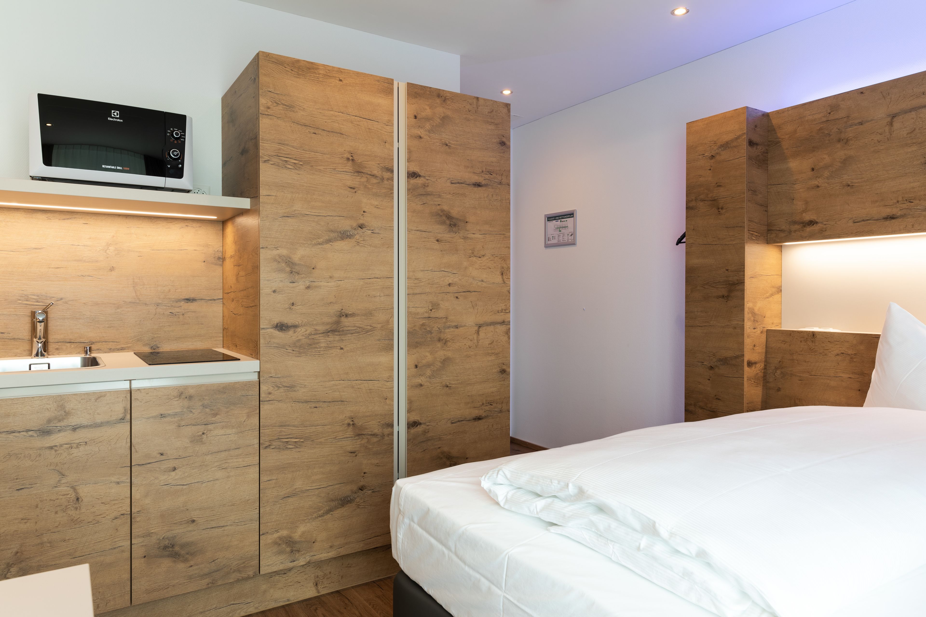 Een moderne slaapkamer met een houten garderobe, een ingebouwde magnetron, een nachtkastje en een bed met wit beddengoed.