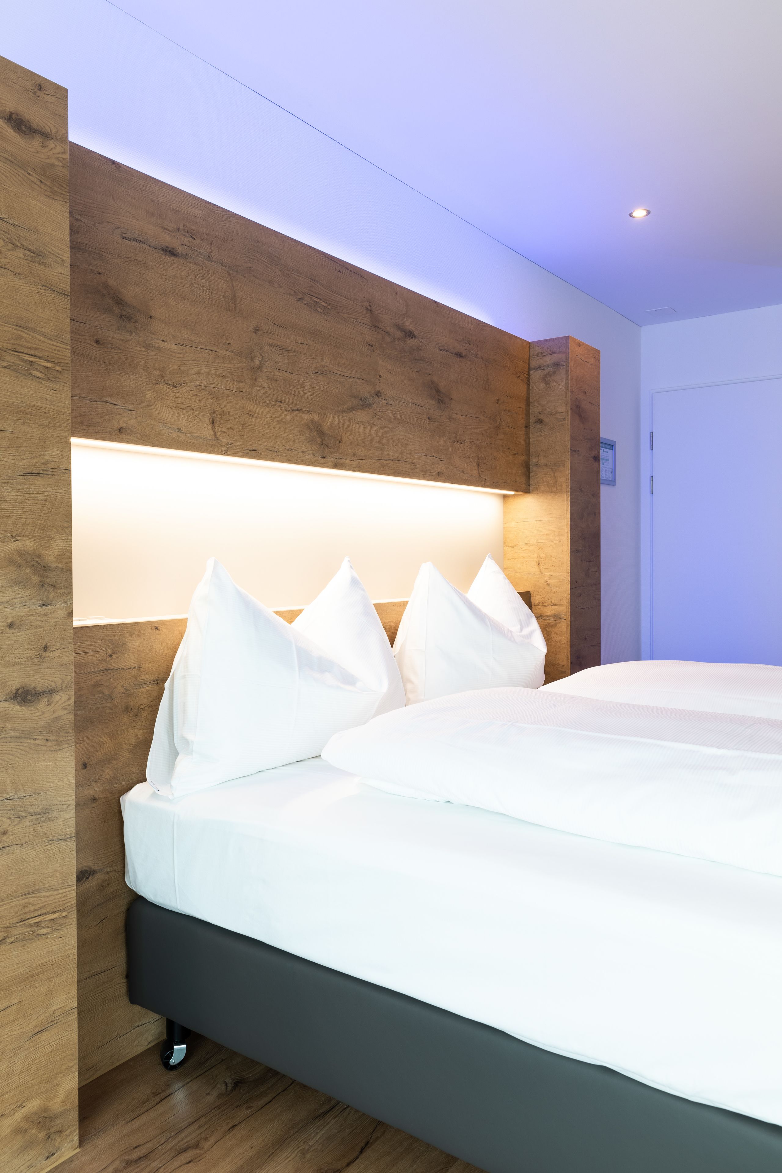 Una camera d'albergo moderna con una testiera in legno, un letto grande con biancheria bianca e una lampada a sospensione luminosa.