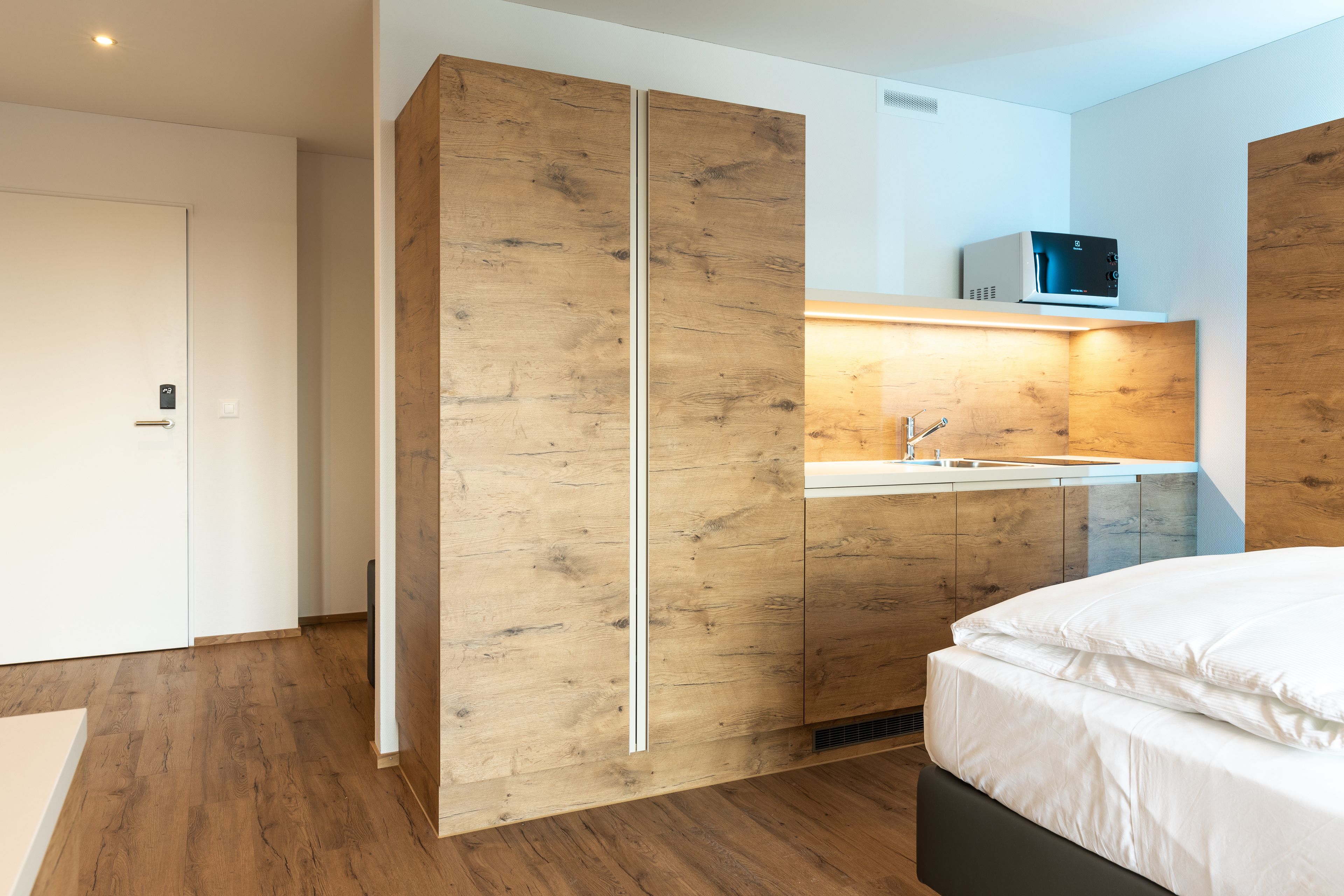 A modern bedroom with a wooden wardrobe, a bed, and a built-in microwave above the sink.