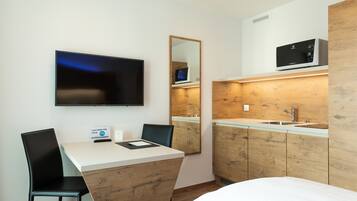 A modern hotel room with a flat-screen TV, a wooden accent wall, a small kitchenette with a microwave, and a desk with a chair.