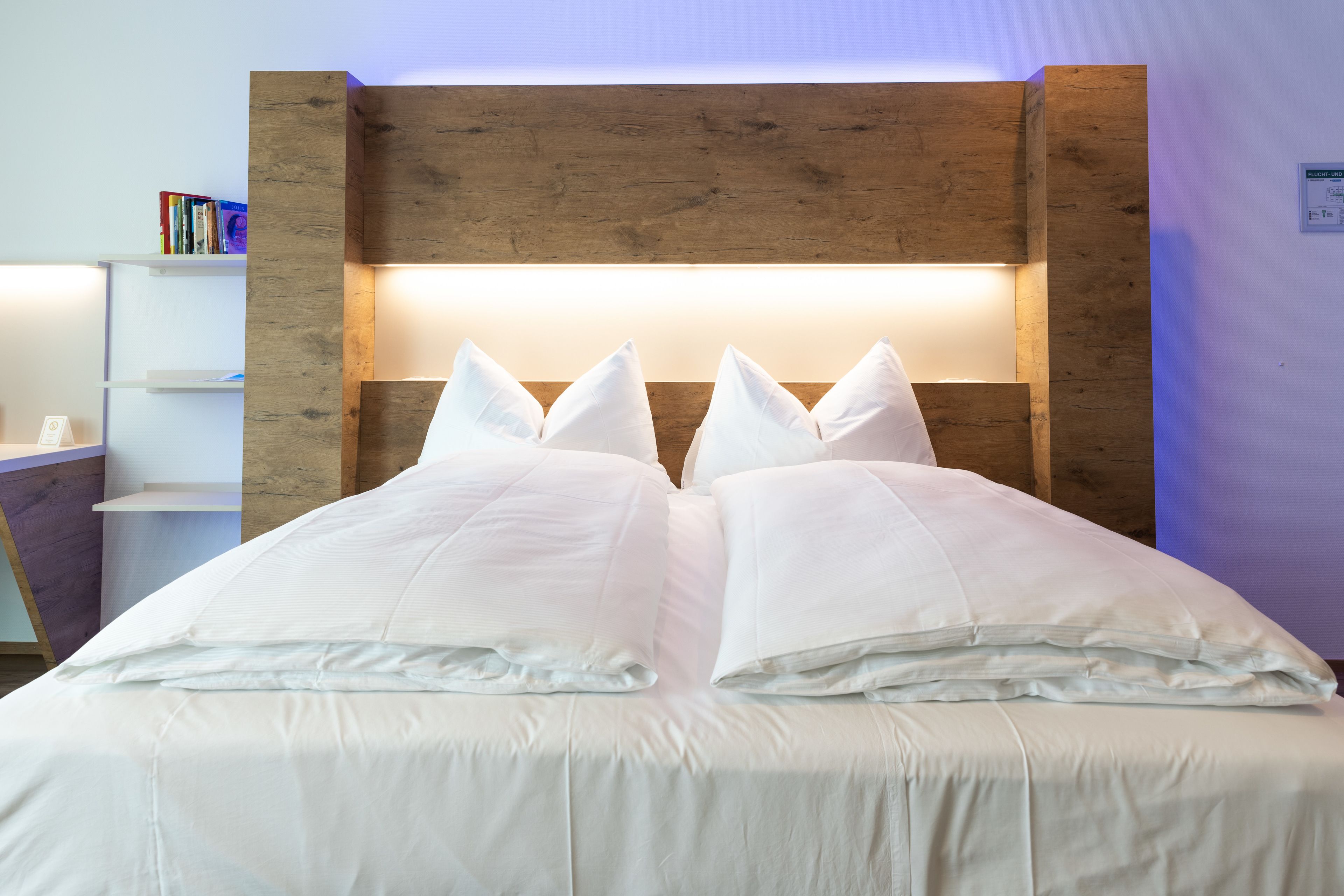 A neatly made bed with white bedding and pillows, a wooden headboard, and a white nightstand with books.