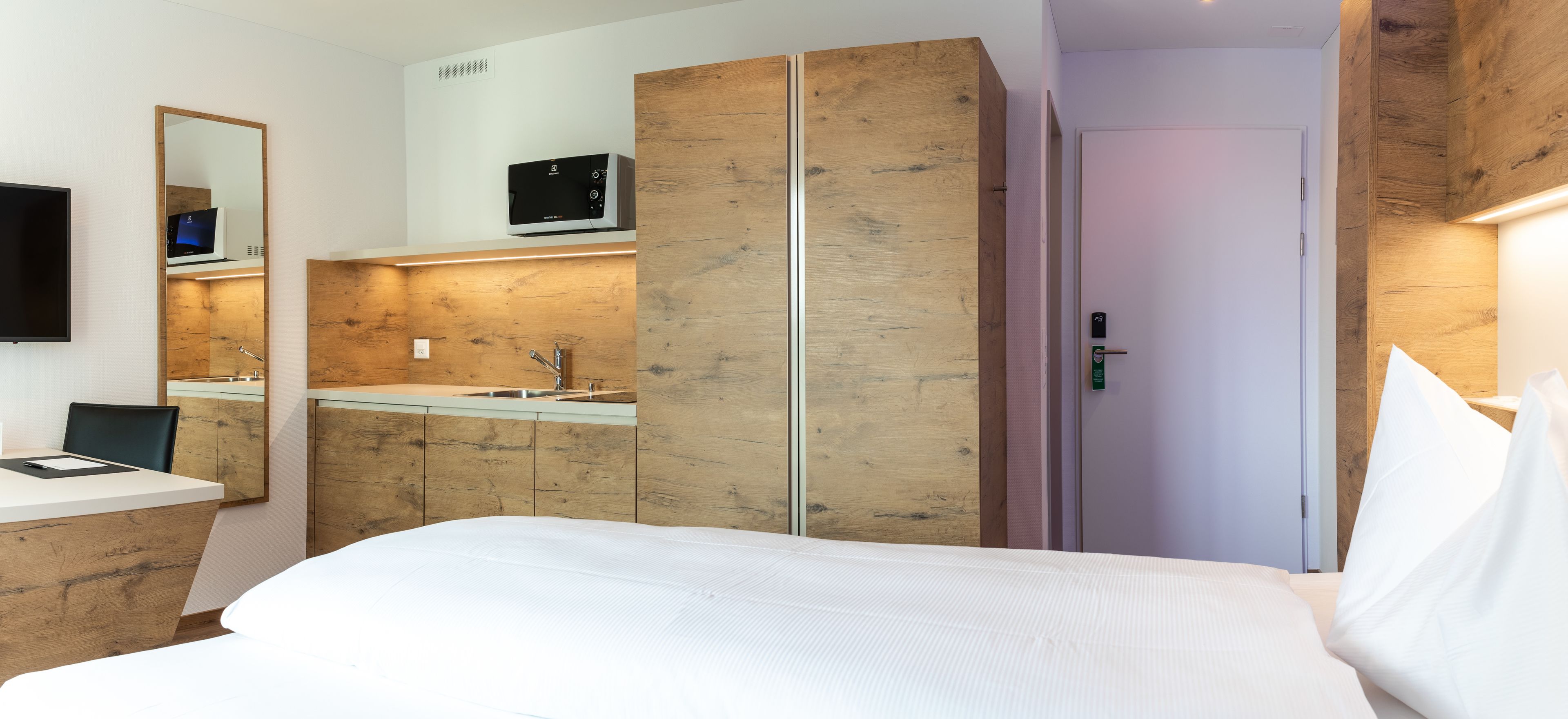 A modern hotel room with a wooden accent wall, a built-in desk, a flat-screen TV, a microwave, and a door with a keycard lock.