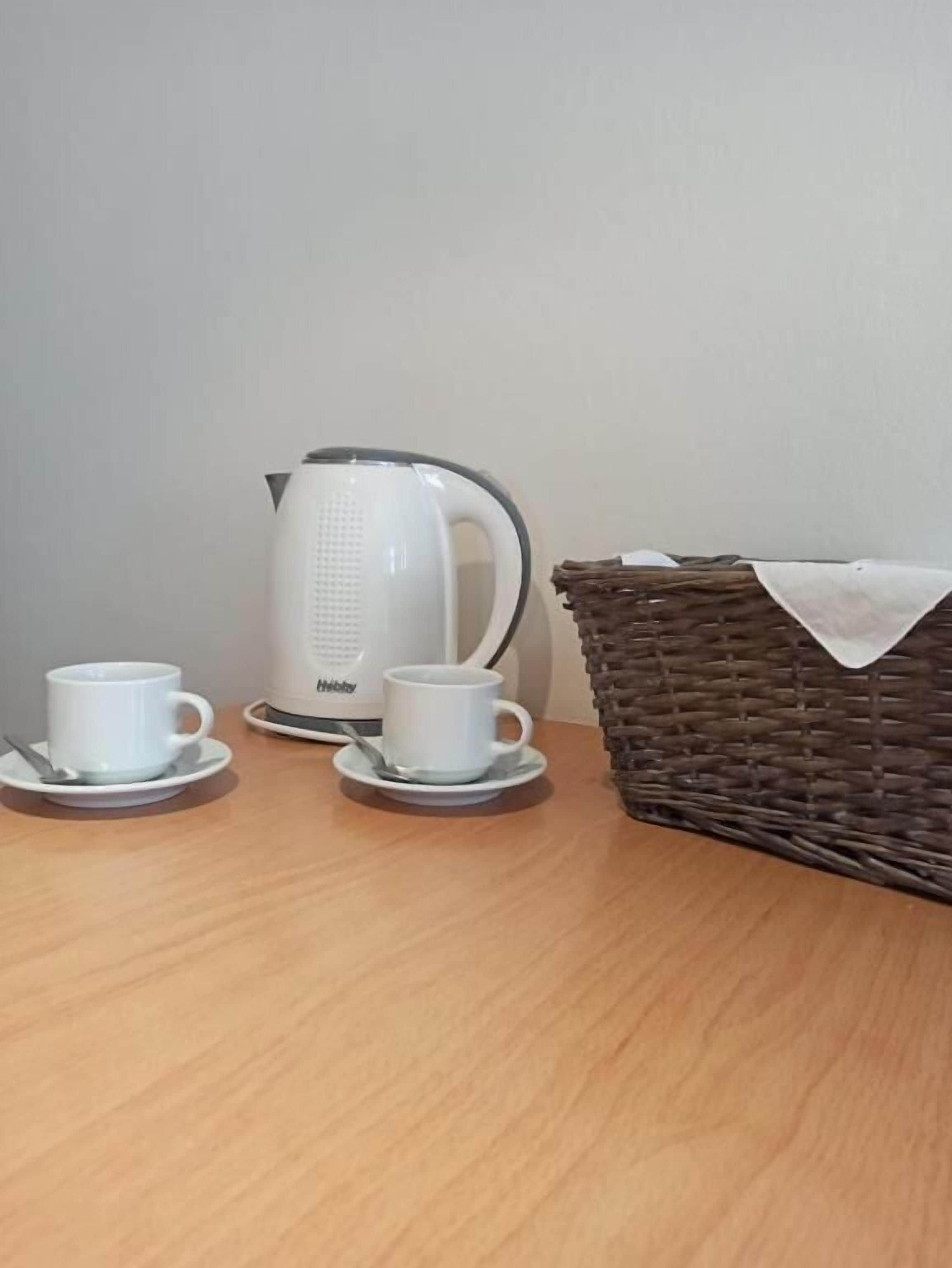 Una mesa de madera con un hervidor blanco, dos tazas blancas y una cesta de mimbre.