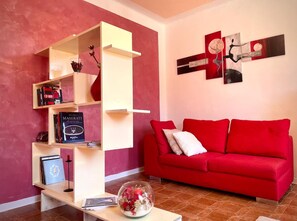 A living room with a red sofa, a white shelving unit, and a glass bowl with flowers on a table. - B&B La Valle Dei Vigher (Cavriana)