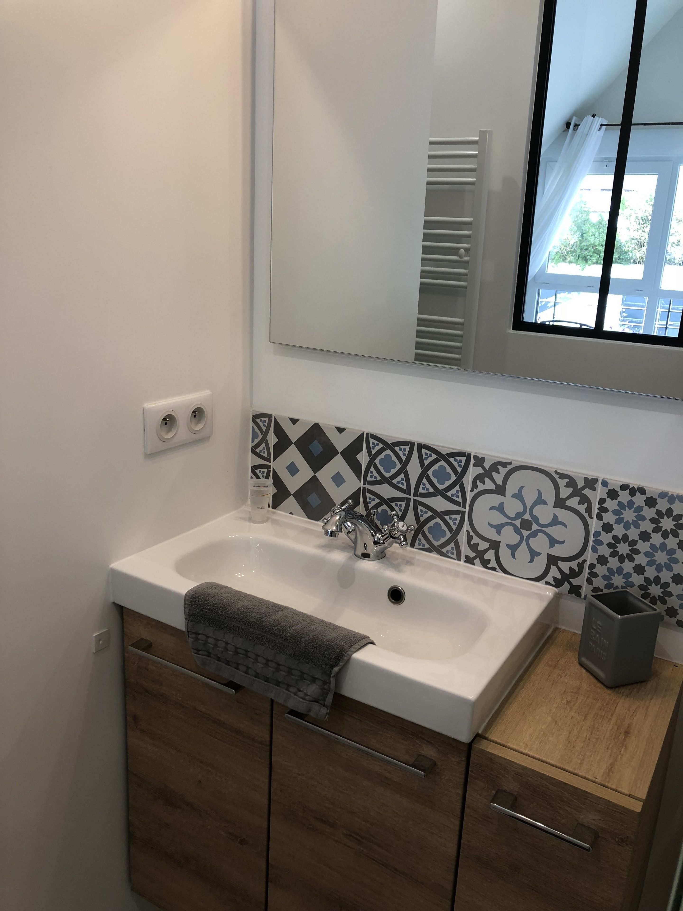 Un baño con un lavamanos blanco, un espejo, baldosas estampadas, una toalla y un cesto de basura.