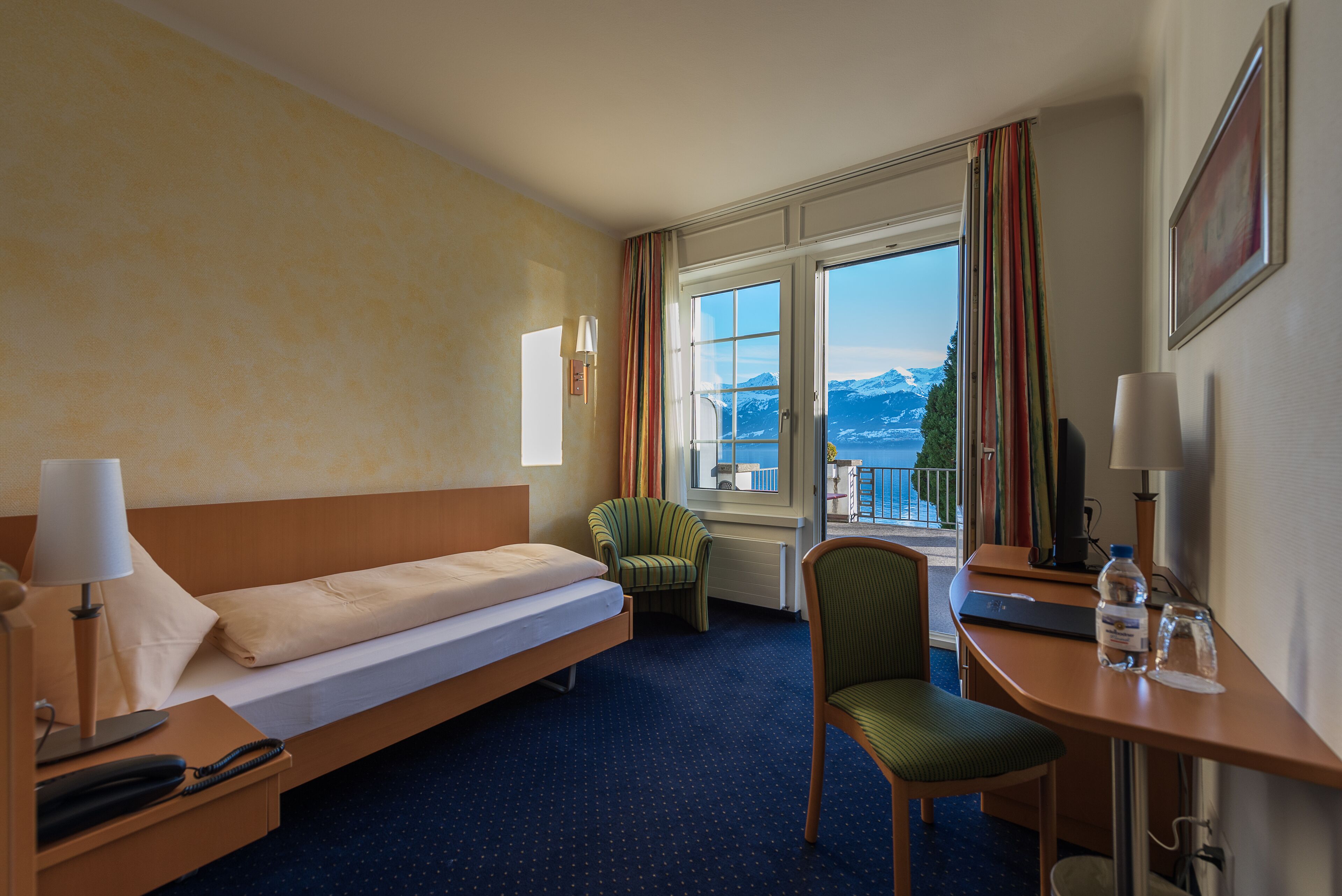 Habitación de hotel con cama, escritorio, silla, ventana y vista a las montañas, además de una lámpara.