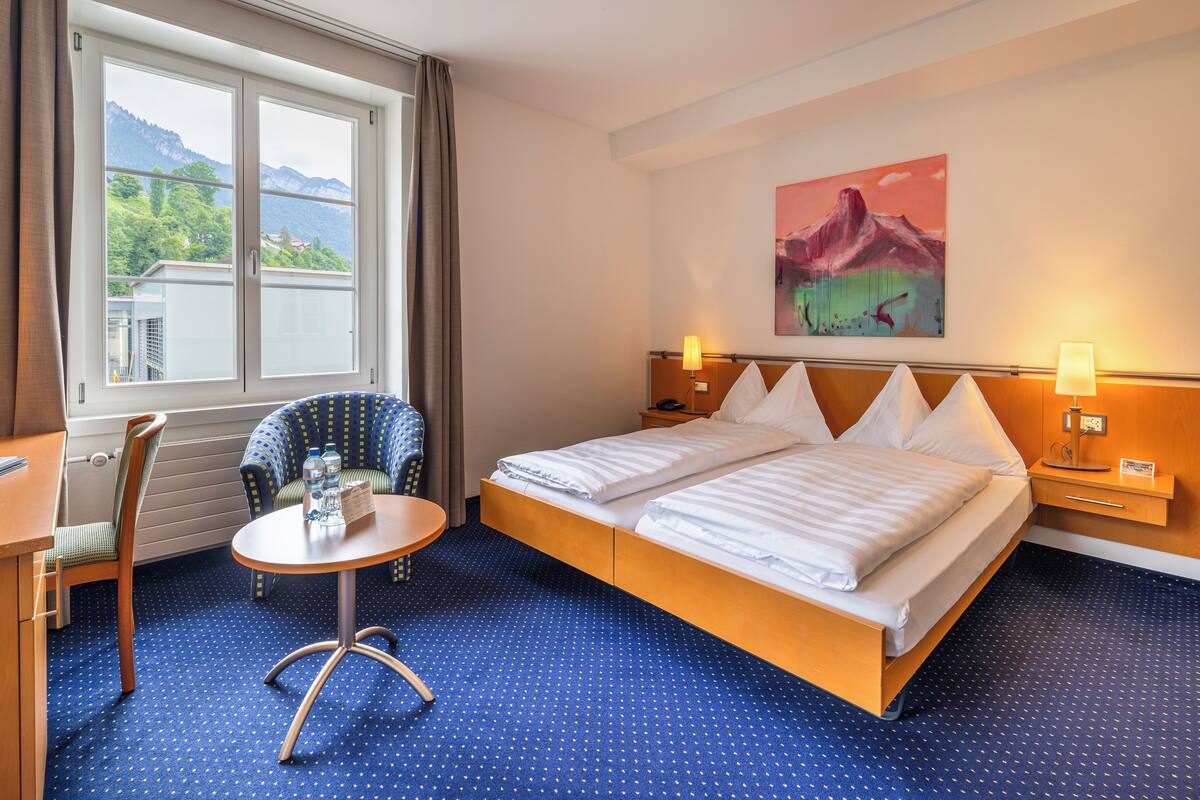 Habitación de hotel con cama, una silla, una mesita y ventana con vistas a las montañas.