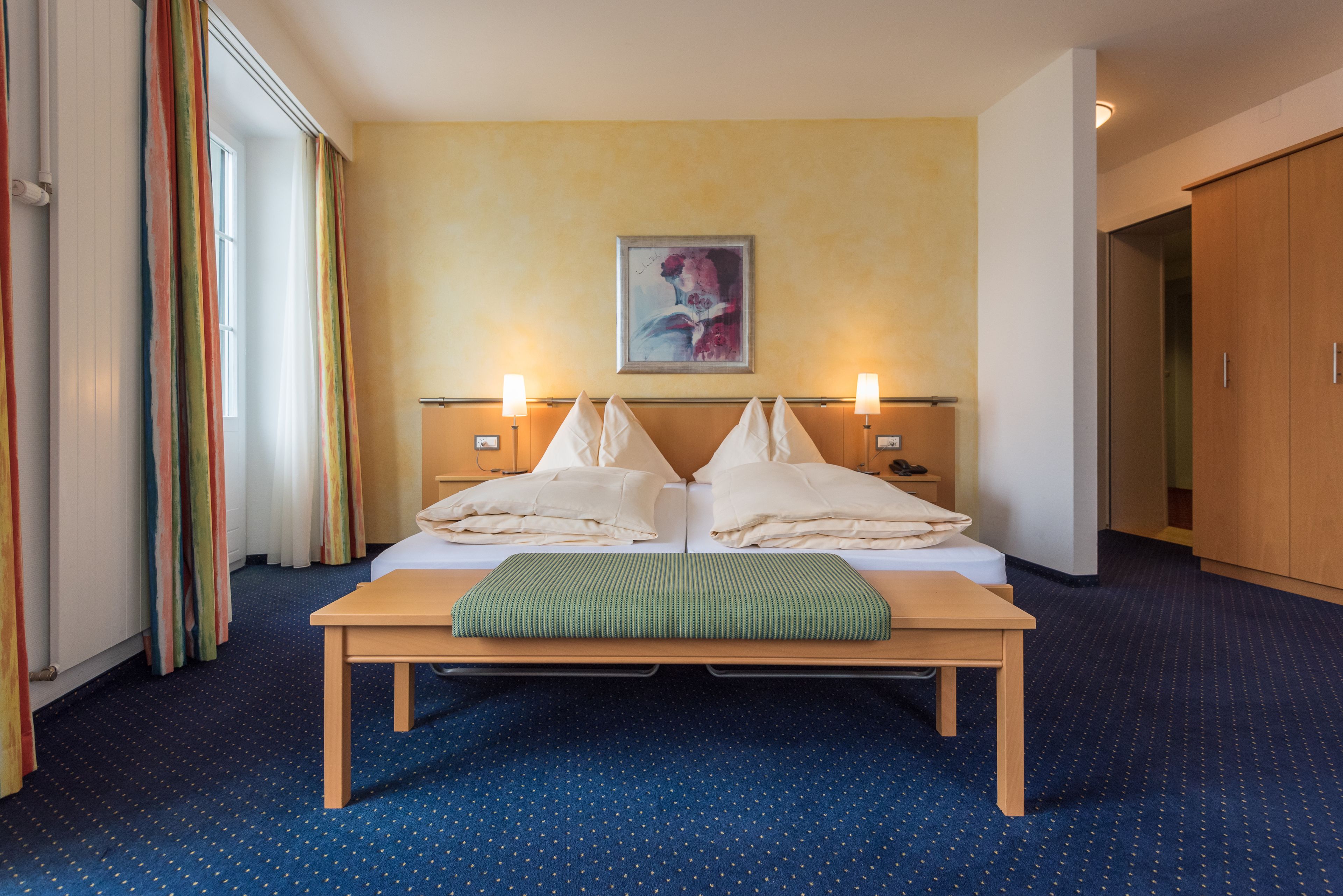 Una habitación de hotel con una cama, lámparas de noche, un cuadro en la pared y un ventanal con cortinas.