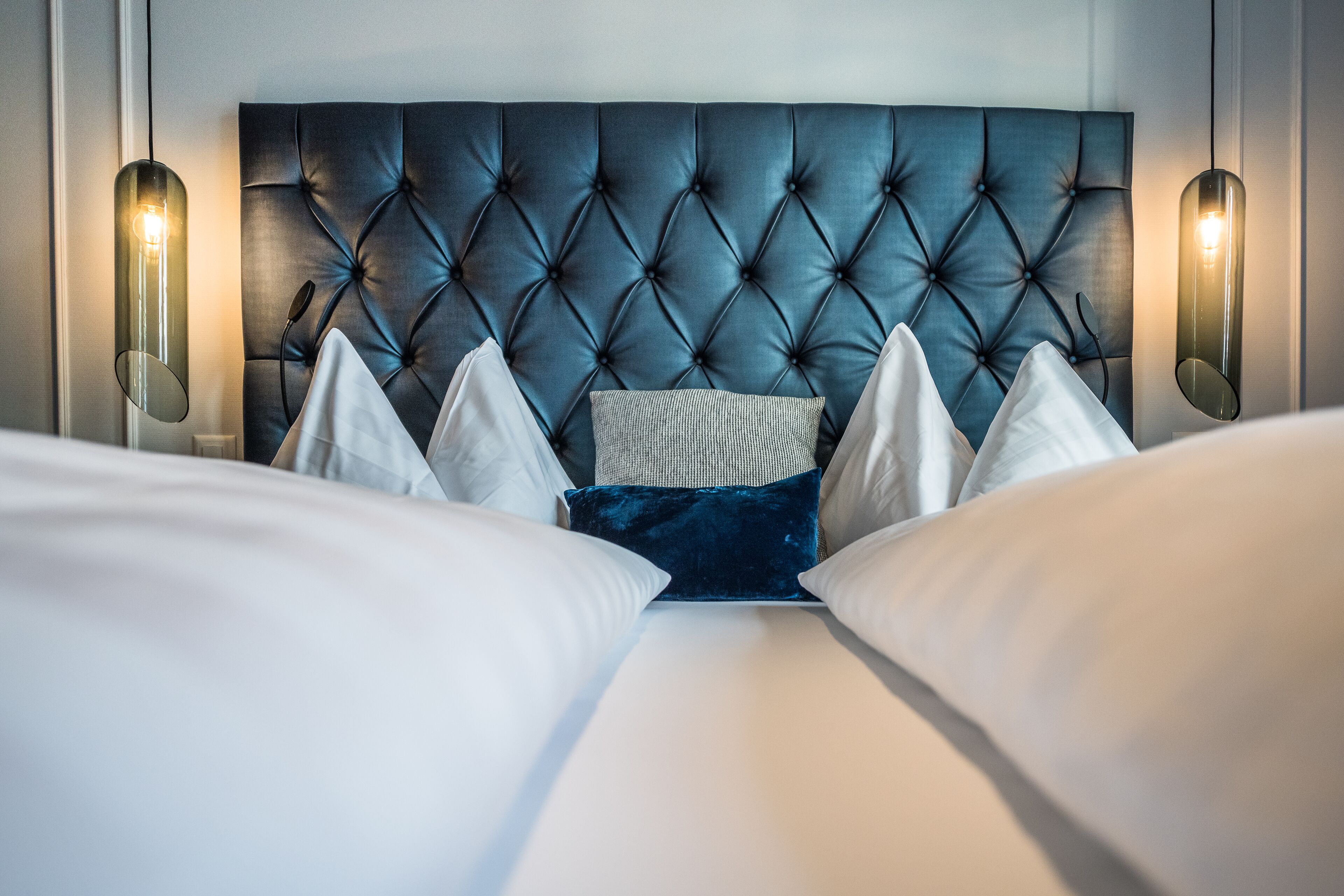 Un lit doté d’une tête de lit capitonnée, de coussins blancs et bleus, et de deux lampes fixées au mur.