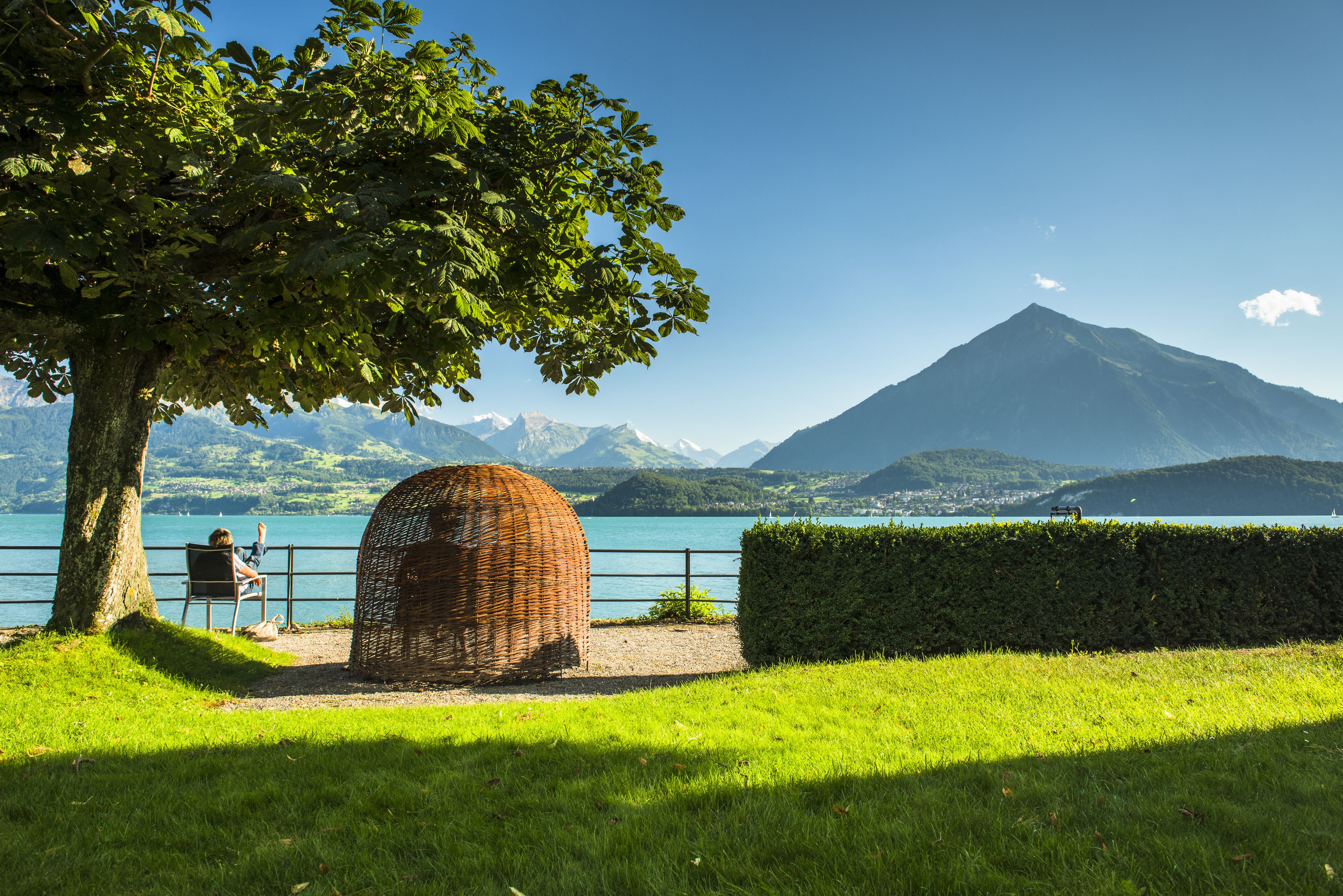 A view of a lake with a mountain in the background, a wicker basket, and a person sitting on a chair.