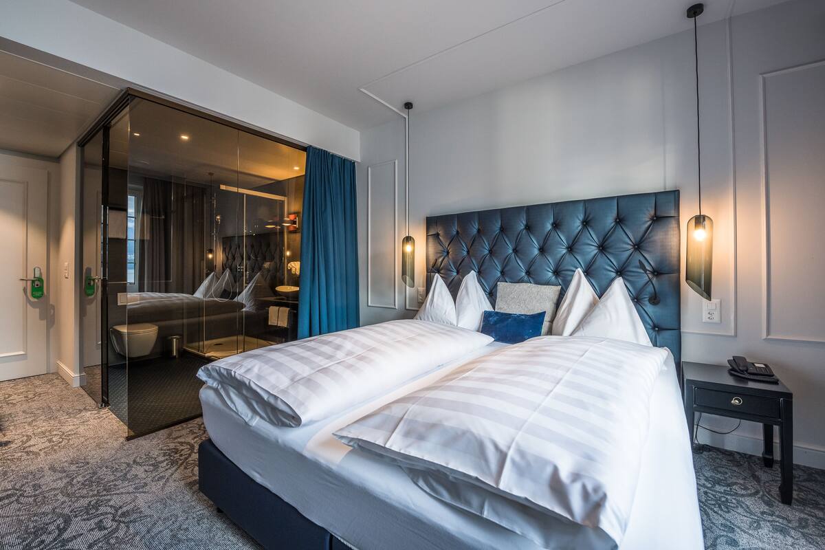 Una habitación de hotel moderna con una cama grande, una puerta de vidrio que da a un baño y un cabecero capitonado.