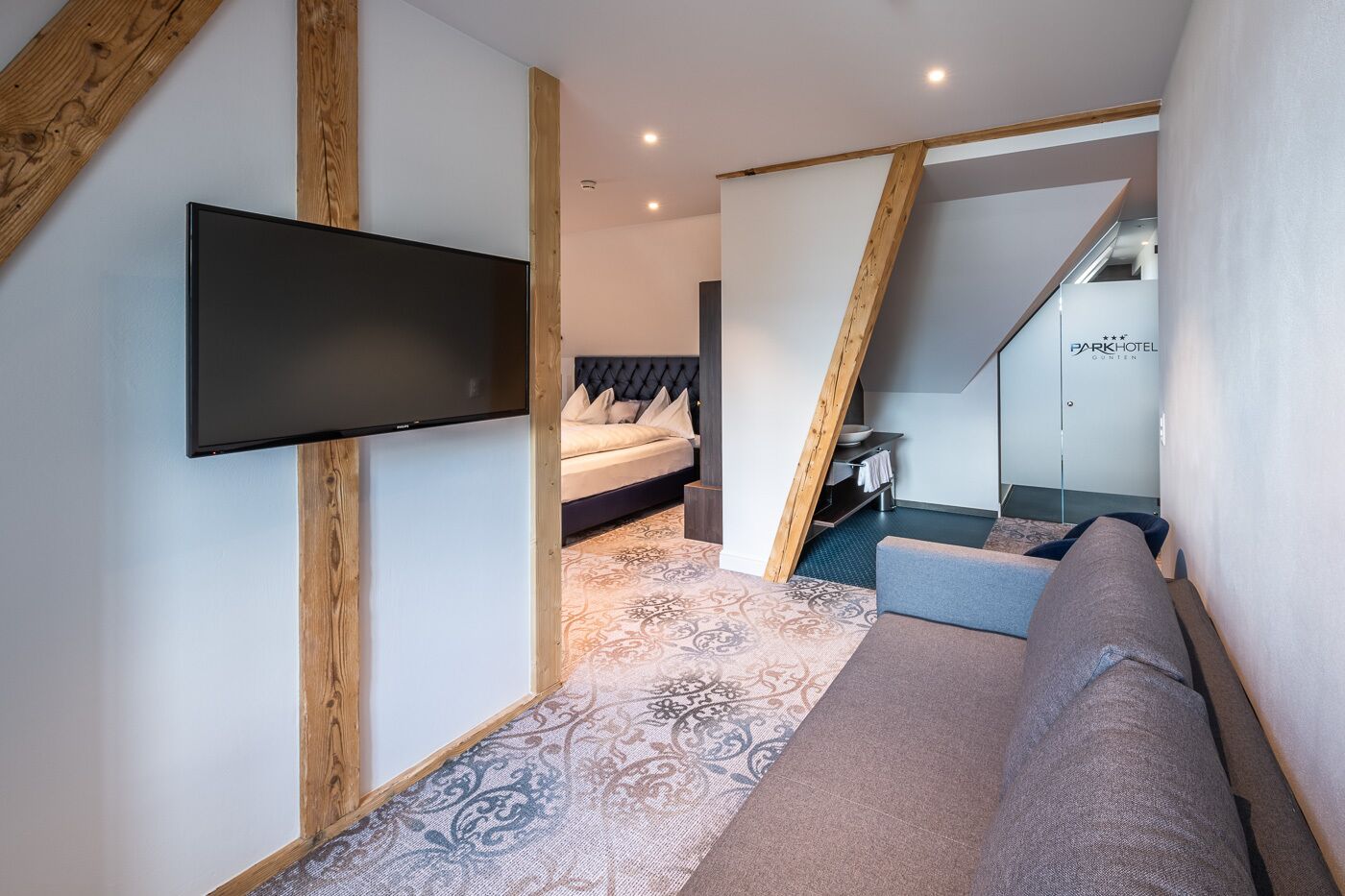 Una habitación de hotel moderna con una gran televisión de pantalla plana montada en la pared, una cama con cabecera oscura, una alfombra estampada y un sofá.