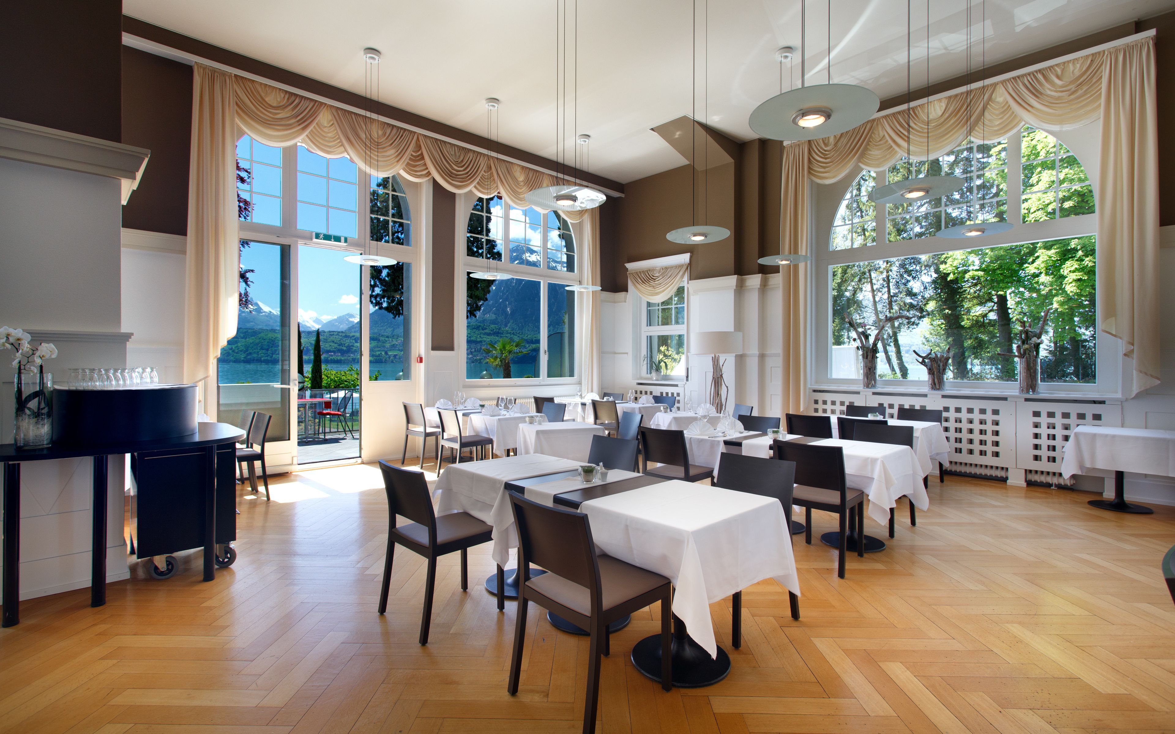 Un comedor amplio con varias mesas y sillas, ventanas grandes que ofrecen vistas panorámicas y una lámpara moderna.