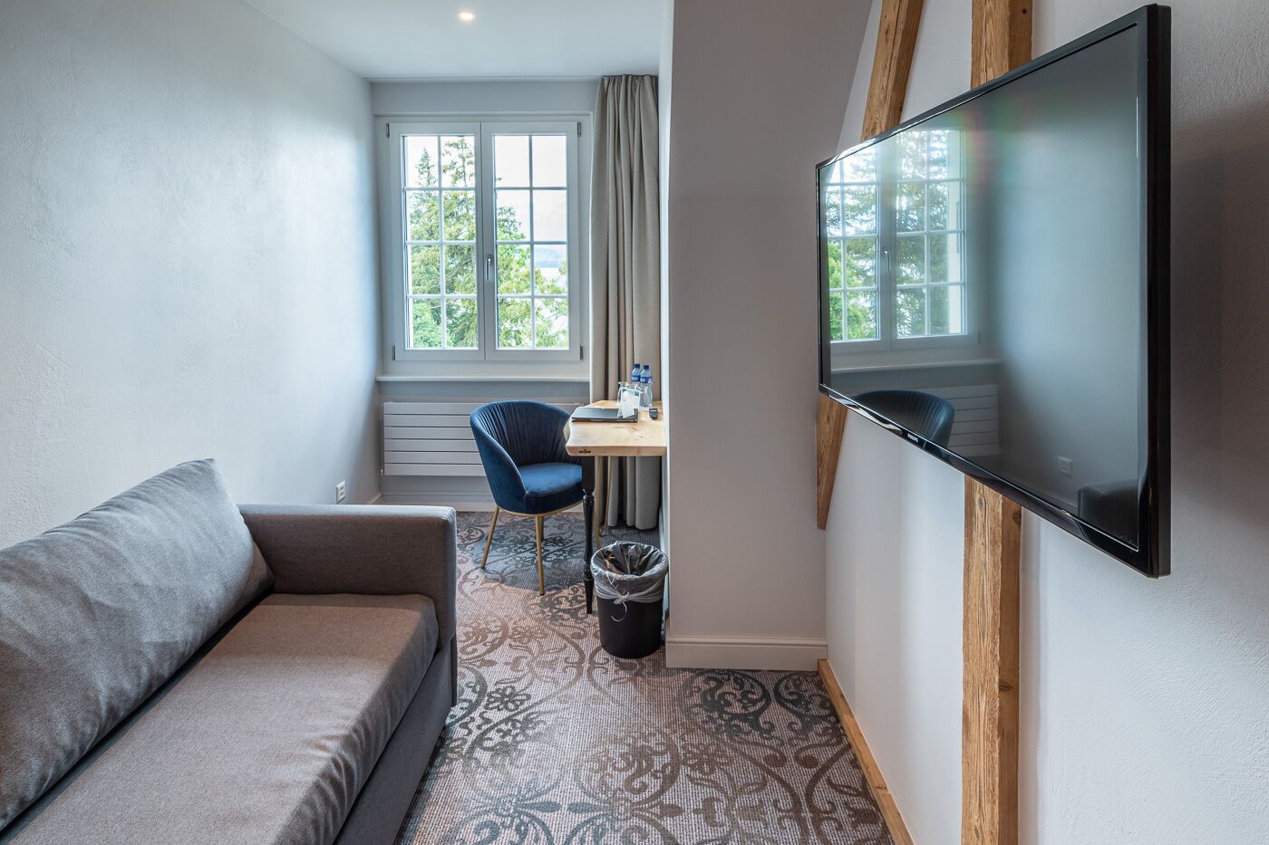 A compact hotel room with a sofa, a small desk with a chair, a television mounted on the wall, and a window with curtains.